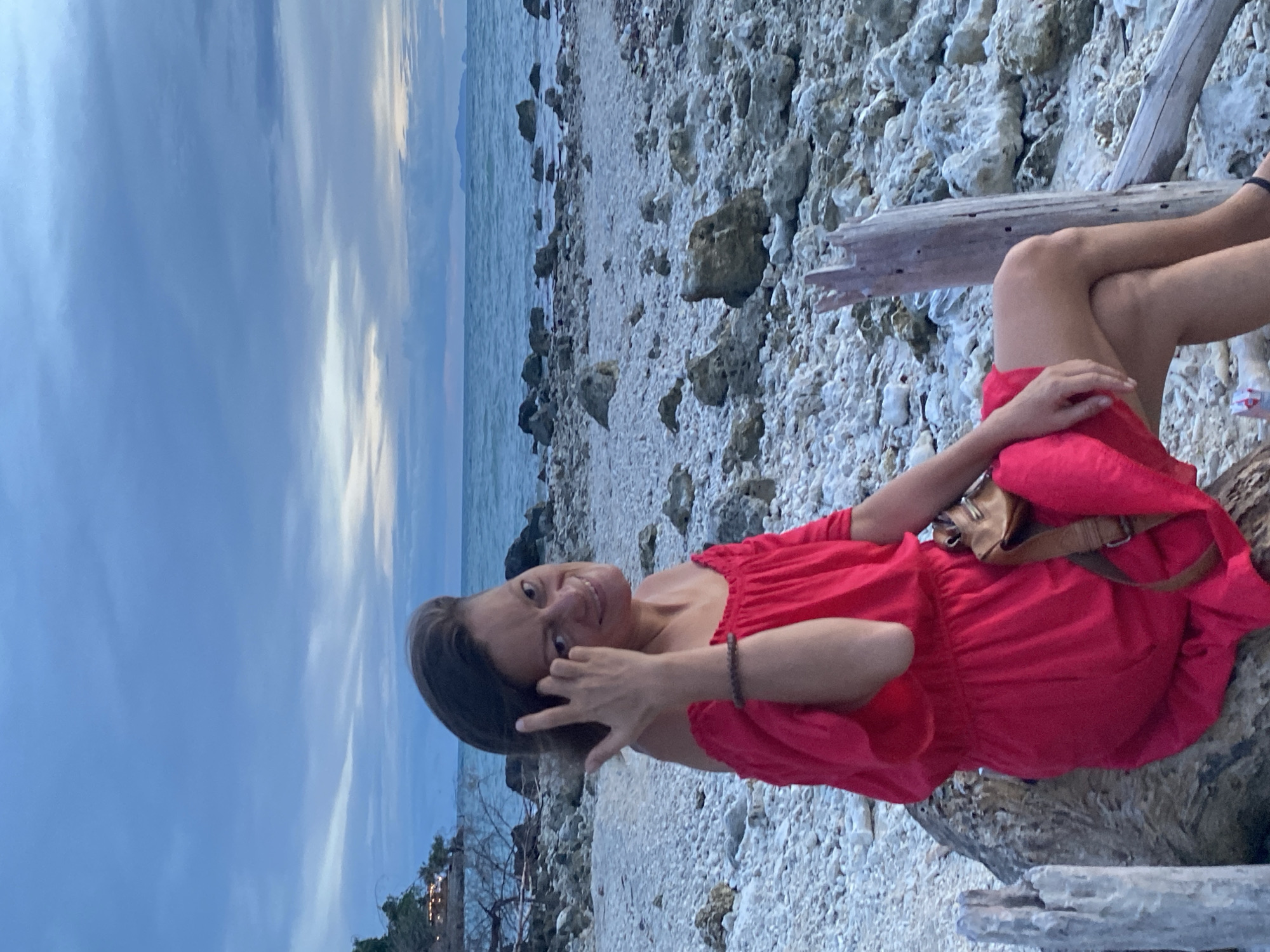 A woman in a bright red off-the-shoulder dress is sitting on a large rock on a pebble beach. She is smiling and looking at the camera, with her right hand touching her hair. A brown crossbody bag is slung over her shoulder. The background shows the ocean and a cloudy sky. The beach is covered in white pebbles and scattered rocks.