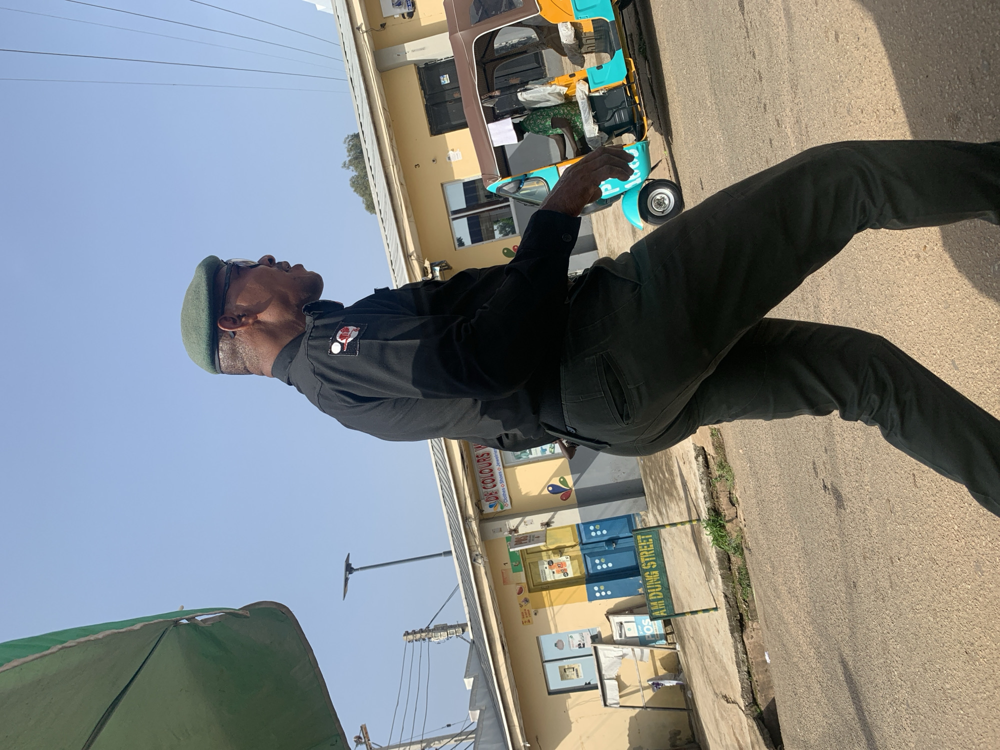A man in a dark uniform with a green beret and glasses walks on a paved street in Jos, Nigeria. He is wearing a black long-sleeved shirt with patches on the left sleeve and dark pants. Behind him, a yellow and blue building has signs that read "DE COLOURS" and "AM DUNG STREET." A turquoise and yellow auto-rickshaw with a driver is parked on the right side of the street. The sky is clear and blue, and a green umbrella is visible in the upper left corner of the image.