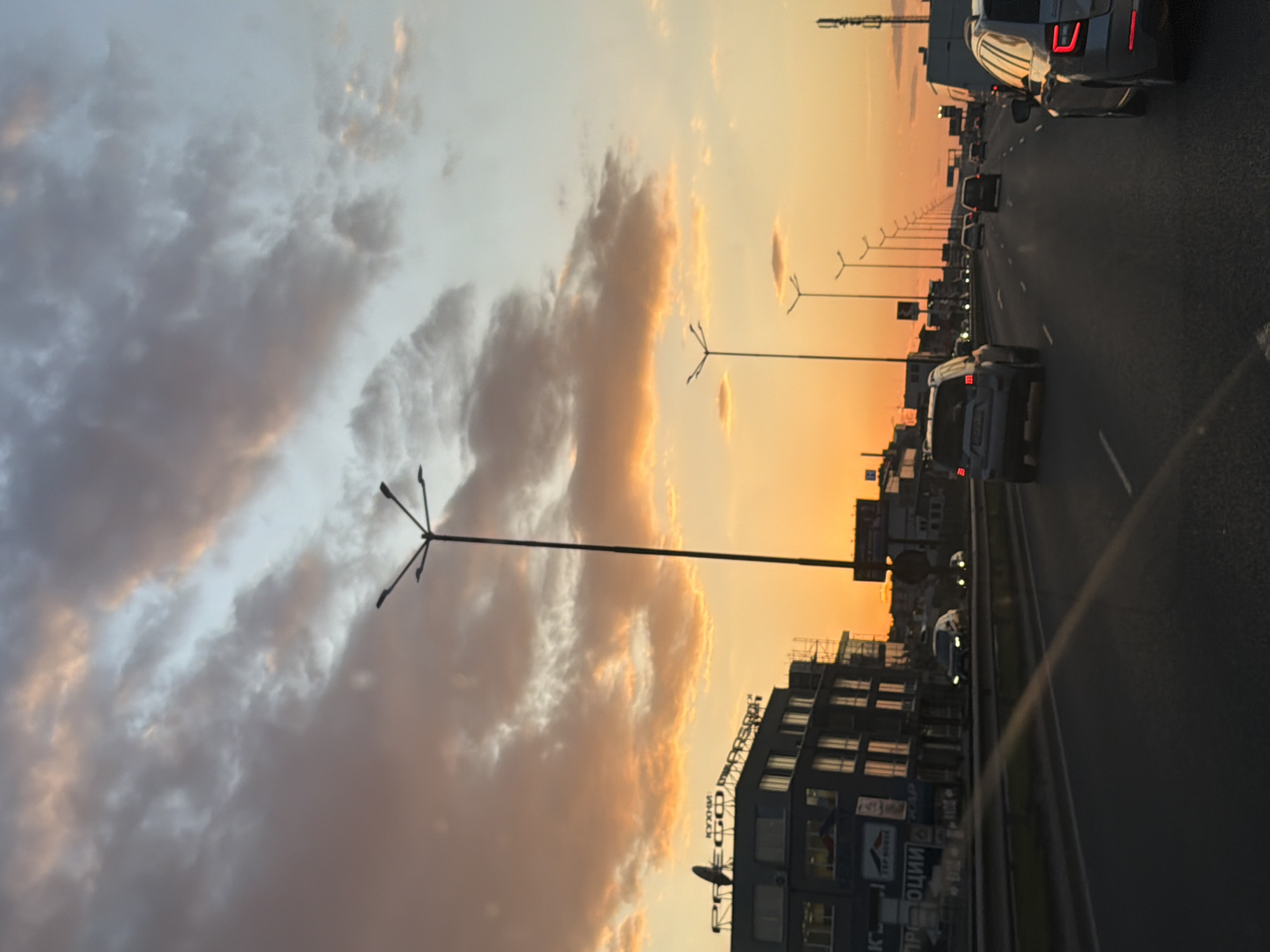 The image depicts a city street or highway at either sunrise or sunset, creating a dramatic, warm-toned sky. The scene is oriented in landscape, with the sky dominating the upper portion and the road and buildings filling the lower half.
In the sky, large, textured clouds are illuminated by the low sun. The color transitions from a vibrant golden-orange on the right side, where the sun appears to be, to cooler grey-blue and light grey tones on the left. The clouds themselves show varying shades of orange, pink, and grey, reflecting the twilight conditions.
Below the sky, a multi-lane road extends into the distance, filled with numerous cars. Many vehicles have their tail lights illuminated, suggesting either slow-moving traffic or a stopped queue. The road surface is dark, with visible white lane markings. Tall, dark streetlights with multiple arms line both sides of the road, receding into the background.
On the left side of the road, a series of modern, multi-story buildings are visible. These buildings feature numerous windows and some display commercial signage.
Visible text on the building signs includes "КУХНИ" (KUKHNI - meaning "Kitchens") and "MEGO" on one sign, and "ΤΕΡΡ НOUSE" (TERR HOUSE) on another. Fragments of other Cyrillic text, including "ШОП" (SHOP) and "...ОЦИИ", are also discernible on various signs.
The overall scene captures a busy urban environment during a picturesque twilight, with the warm hues of the sky contrasting with the darker tones of the city infrastructure and vehicles. The location is Sofia, Bulgaria.