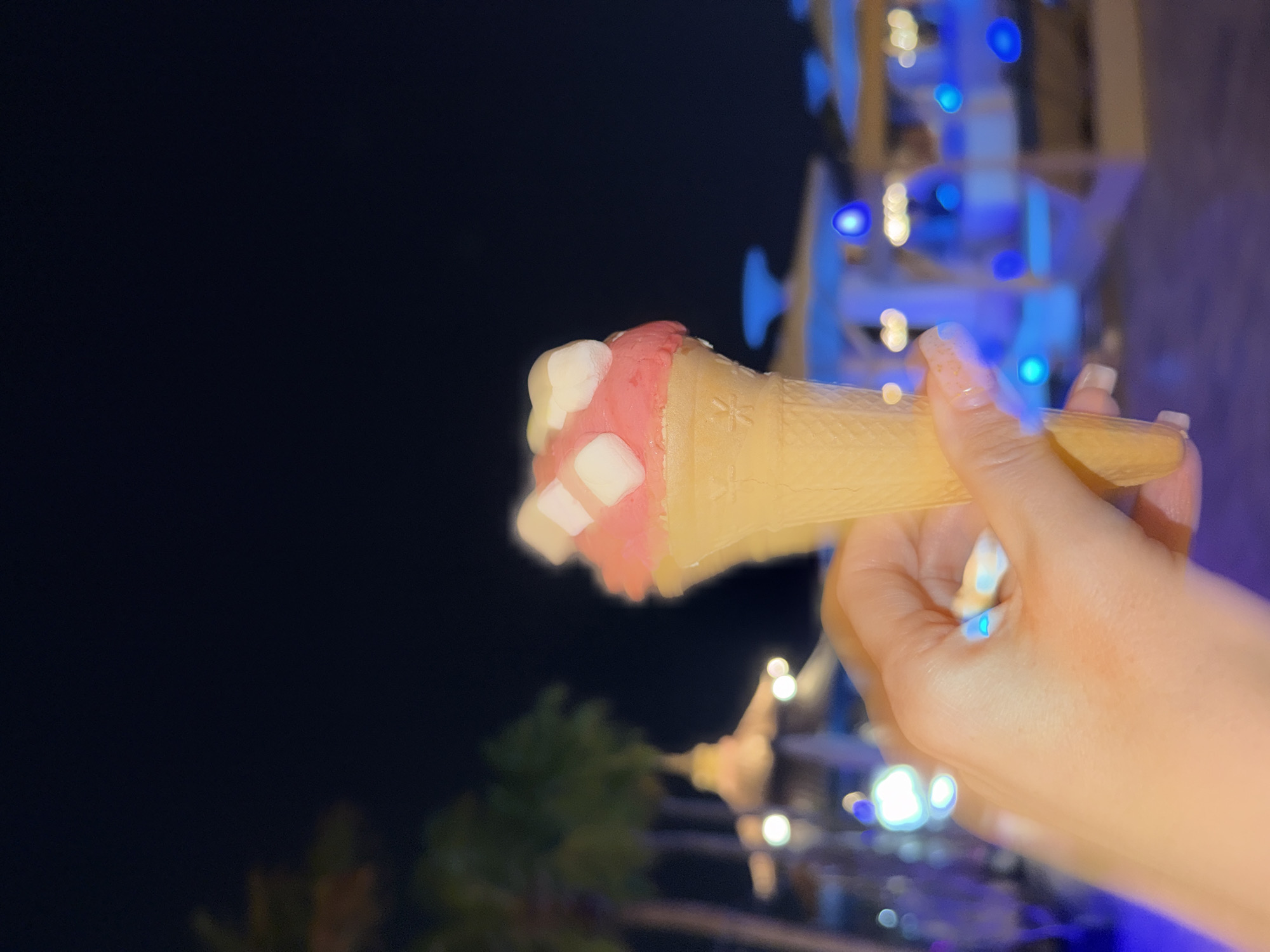 A close-up, low-angle shot captures a hand holding a pink ice cream cone topped with white cube-shaped marshmallows against a blurred nighttime background. The hand is light-skinned with a light pink manicure and appears to be positioned in the foreground. The ice cream cone itself is light yellow and waffle-textured. The background is a dark blue night sky with out-of-focus elements that suggest a vibrant city environment, possibly a resort or entertainment district in Dubai, United Arab Emirates, indicated by the presence of palm trees and illuminated structures with blue and white lights. The overall impression is of a casual, nighttime scene.