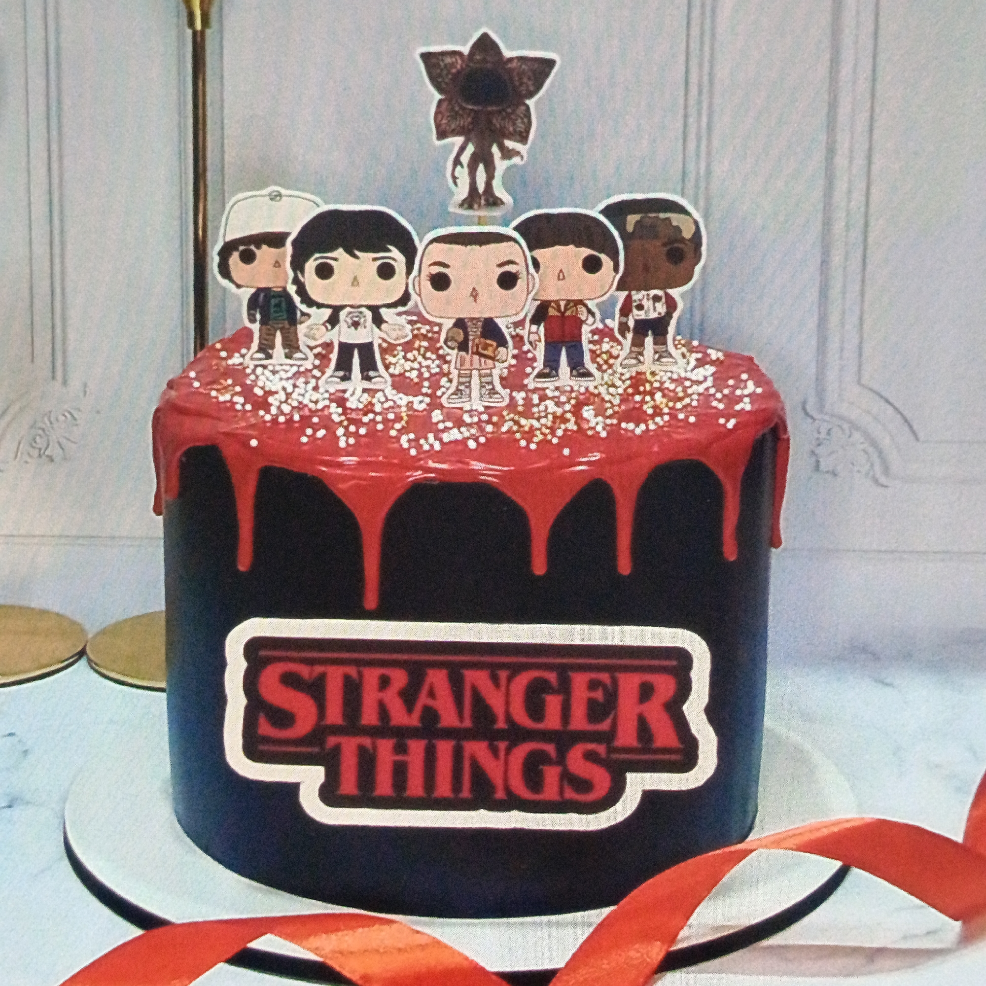 A decorated cake is positioned in front of a wall panel that appears to be painted in a light color. The cake is black with a red topping drizzled down the sides and sprinkled with small white decorations. The front of the cake has a "Stranger Things" logo. On top of the cake are paper cutouts of "Stranger Things" characters. A red ribbon is draped in the foreground. The cake is located in the city of Марковский, Russia.