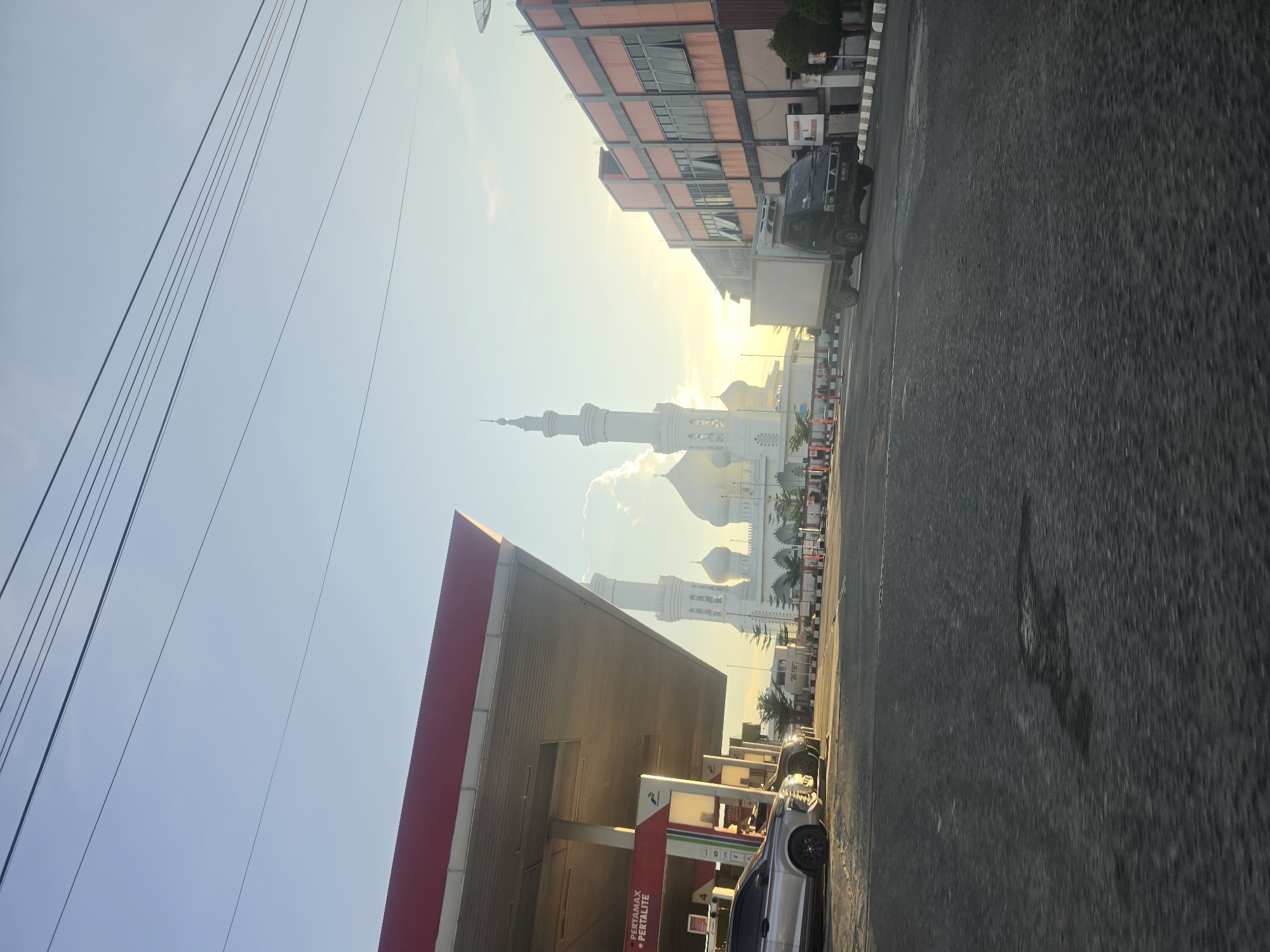 The image, taken in Ranto, Indonesia, presents a street scene with a mosque visible in the background. The foreground includes a gas station with a "PERTAMAX PERTALITE" sign. A silver car is parked at one of the pumps. On the opposite side of the street, there is a multi-story building. Overhead power lines are visible against a clear sky. A truck and several cars are on the road, facing towards the mosque. The overall lighting suggests it is early morning or late afternoon.