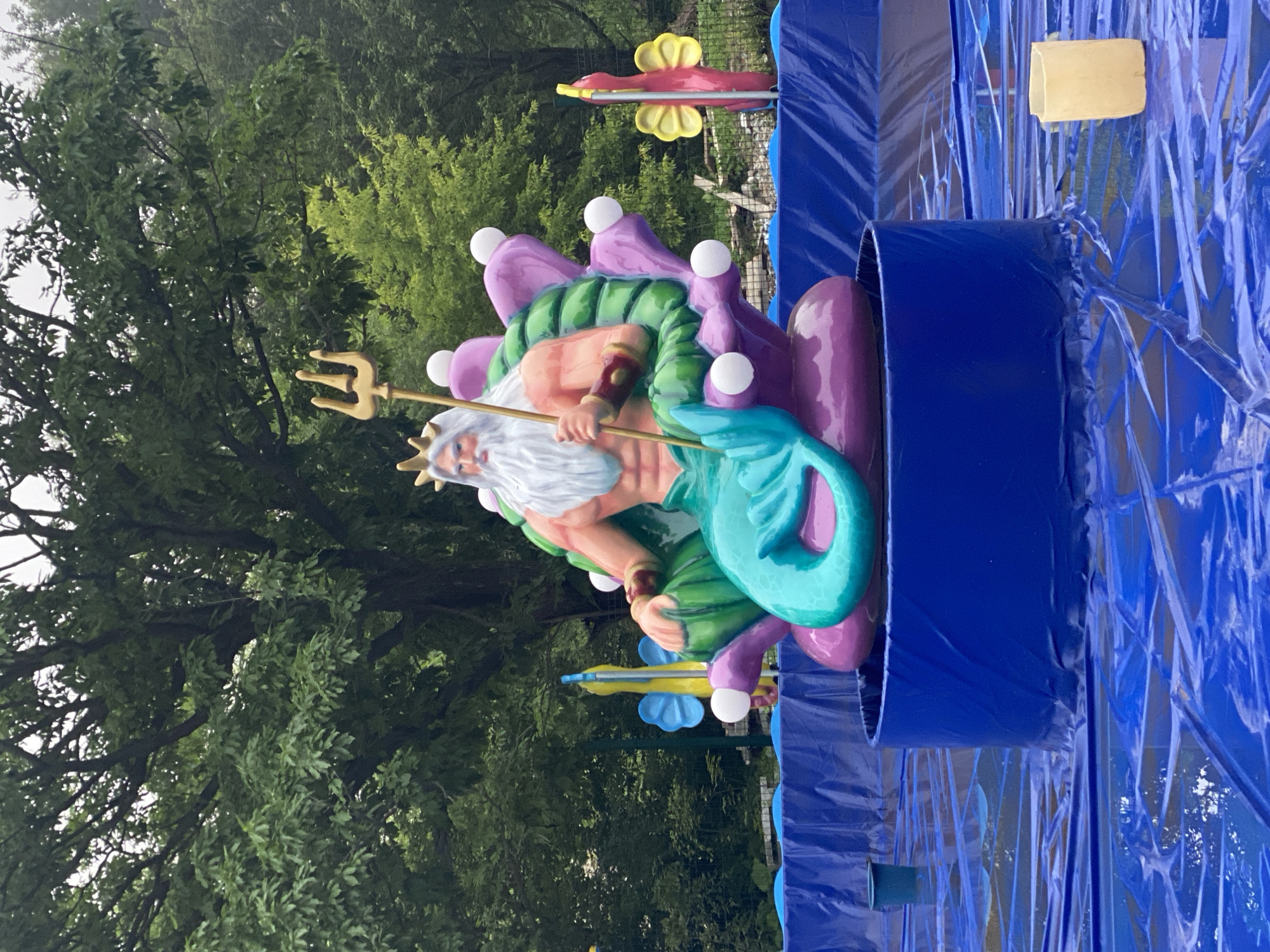 This is a photograph of a statue of Poseidon, the Greek god of the sea, seated on a throne resembling coral. Poseidon is depicted with a white beard, a crown, and a trident in his right hand. He has a muscular, tanned torso and a turquoise mermaid tail. The statue is placed on a blue platform, which is surrounded by blue plastic sheeting that mimics water. In the background, there are trees and two colorful statues of fish. The scene appears to be an outdoor setting, possibly a park or a themed attraction, though the specific location in the city of Unknown, Unknown is not identifiable. The lighting suggests an overcast day.