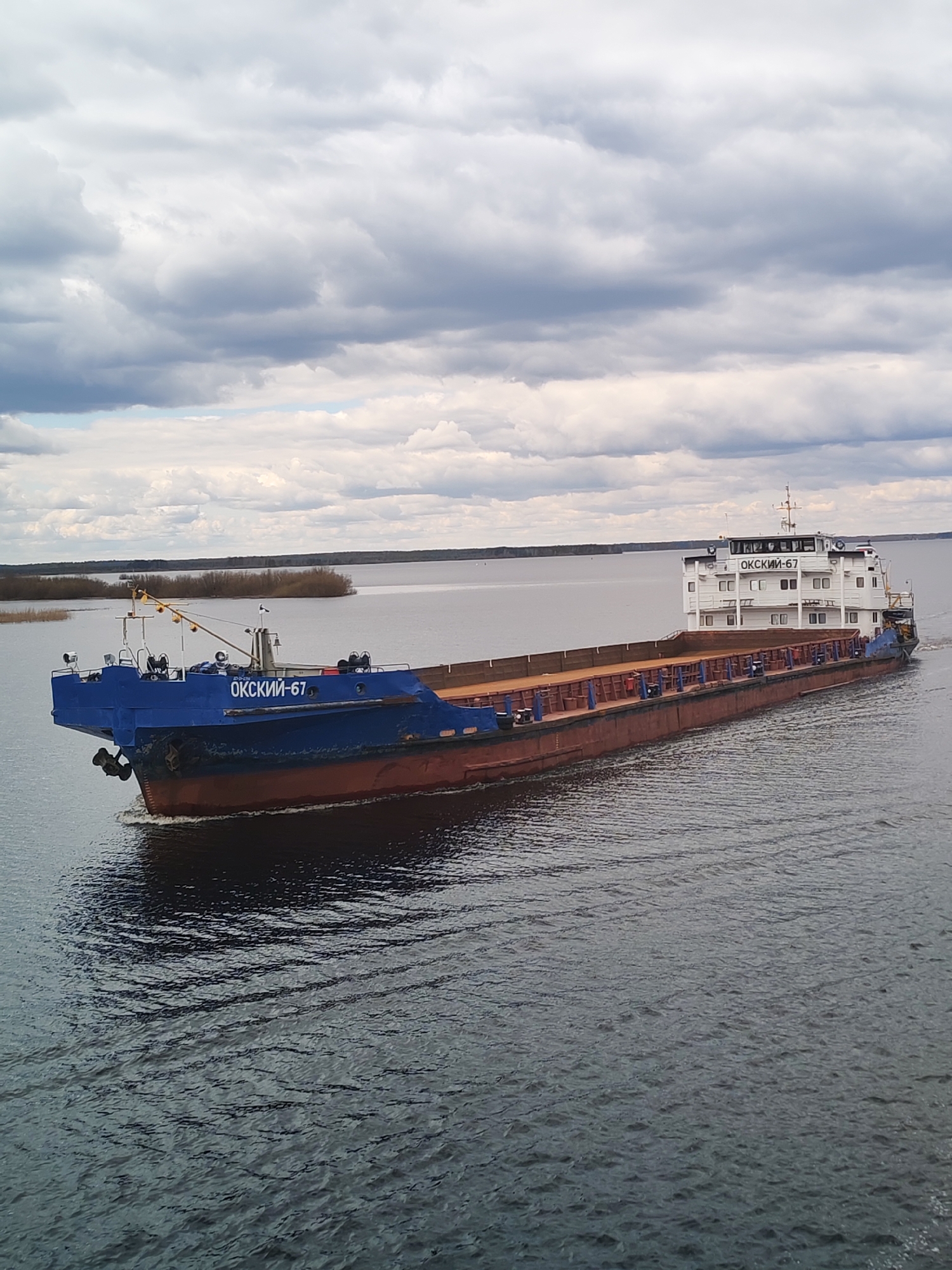 A motor ship named "ОКСКИЙ-67" (OKSKIY-67) is depicted navigating a wide body of water under a heavily overcast sky. The ship's bow is painted blue, featuring the name in white capital letters. Its hull beneath the waterline and along the open cargo deck is a reddish-brown, while the multi-deck superstructure at the stern is white. The vessel is captured in motion, producing a noticeable wake in the dark, rippled water.
In the background, a low, flat shoreline with sparse, muted vegetation is visible in the distance, suggesting a large river or lake, possibly in an early spring or late autumn season. The sky is dominated by thick layers of grey and white clouds, indicating a cloudy day with no direct sunlight. The overall scene conveys a functional and calm environment typical of inland water transport. The name "ОКСКИЙ" (OKSKIY) on the ship likely refers to the Oka River, a major waterway in Russia, consistent with the provided location context of Суда, Russia.