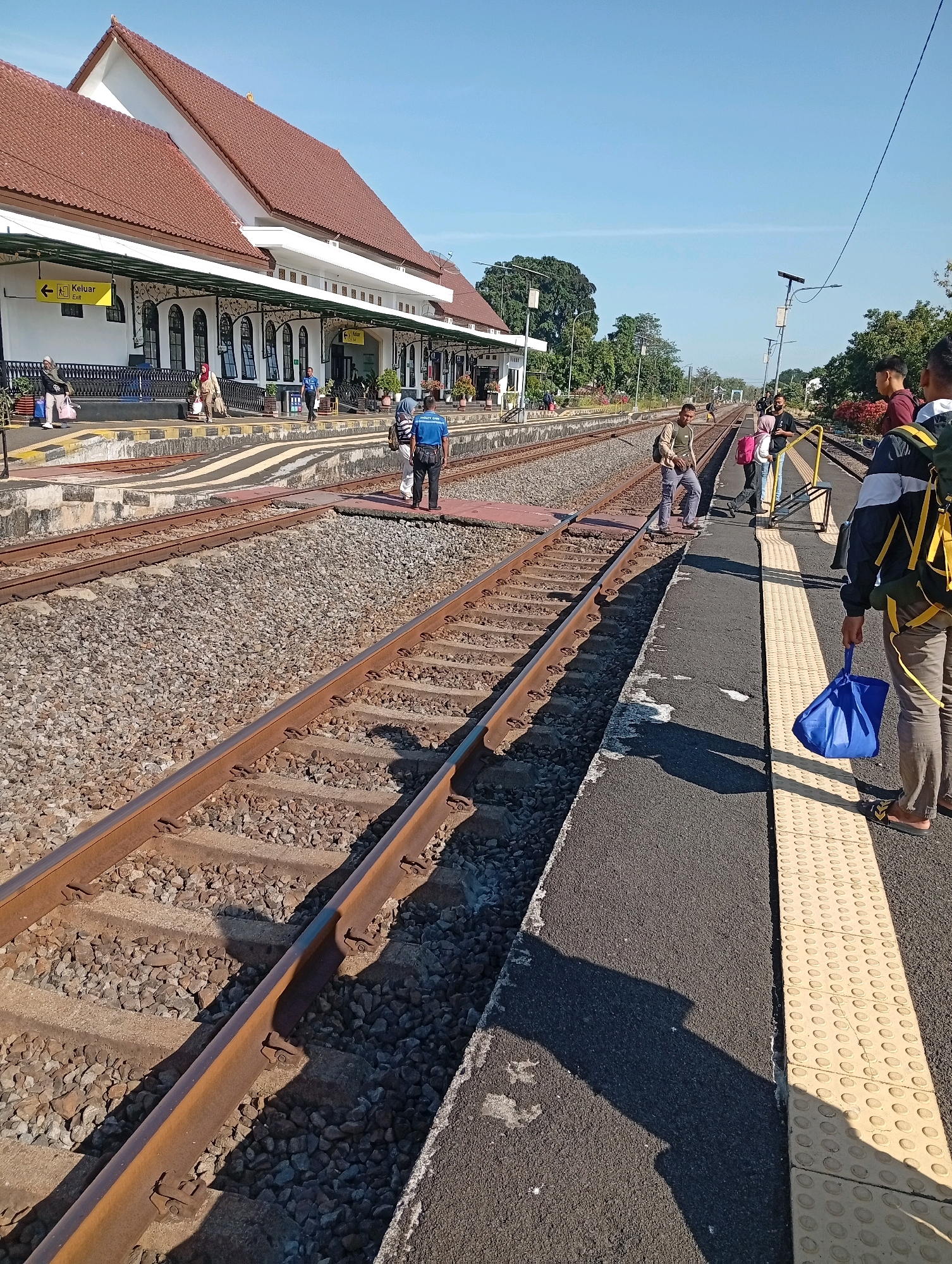 This is a ground-level, slightly angled, full shot of a train station platform and tracks in Tebon, Indonesia. The scene is bathed in bright sunlight, creating distinct shadows. On the left, a long, white building with a red-tiled roof, characteristic of Indonesian colonial architecture, serves as the station. An "Exit" sign with yellow lettering is visible on the building. A train track runs horizontally across the foreground, with another track to its right. Several people are visible on the platform to the right, some with backpacks, suggesting they are travelers. One person in a blue shirt appears to be a station employee. The platform has a textured, yellow tactile paving strip along the edge next to the tracks. The environment is clear and sunny, with trees and vegetation visible in the background.