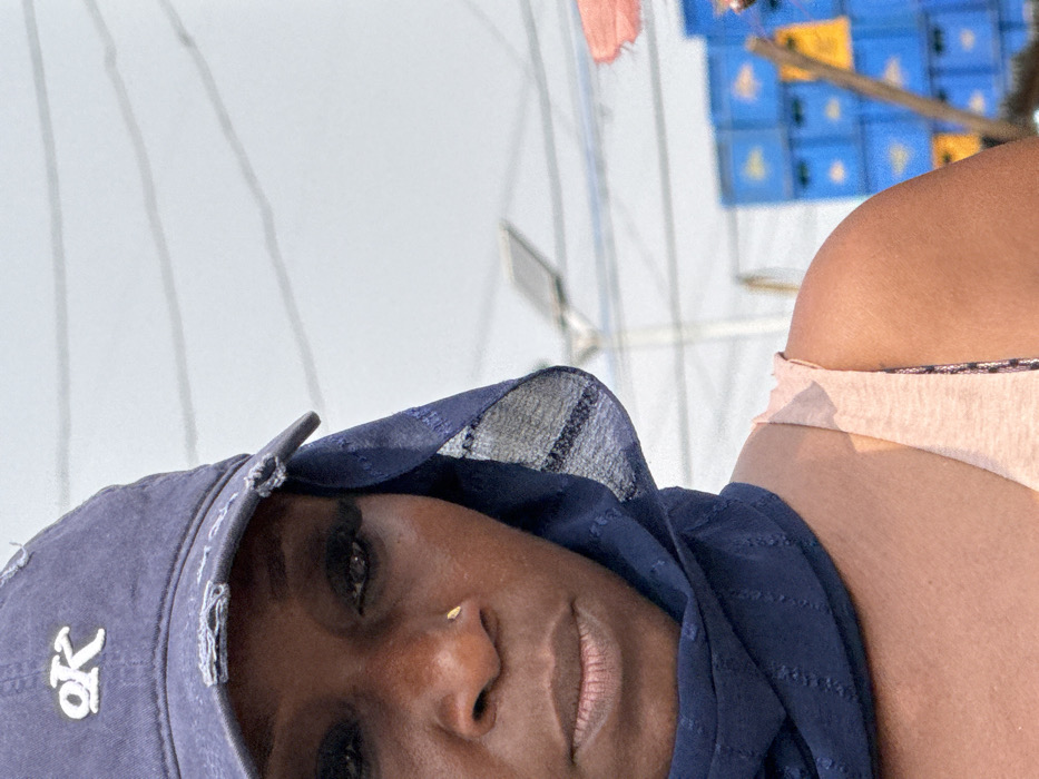 A close-up portrait image captures an adult Black female in an outdoor setting in Jakande, Nigeria. She is positioned on the left side of the frame, facing slightly forward with her head tilted downwards and her gaze directed downwards and to the left. She wears a distressed navy blue baseball cap with a white "OK" logo embroidered on the front, and a dark blue sheer scarf is wrapped around her head and neck. Her facial features include dark eyes with apparent false eyelashes or heavy mascara and a small gold-colored stud in her right nostril. She has a brown skin tone. A light peach-colored sleeveless top is visible on her right shoulder and upper chest. The background is out of focus but shows several horizontal lines, possibly overhead wires, in the upper portion. A pole-mounted street light fixture is visible behind her to the right. Further to the right, a stack of blue plastic crates, some with yellow markings, is present, along with a reddish-orange object, possibly fabric, and a leaning wooden pole.