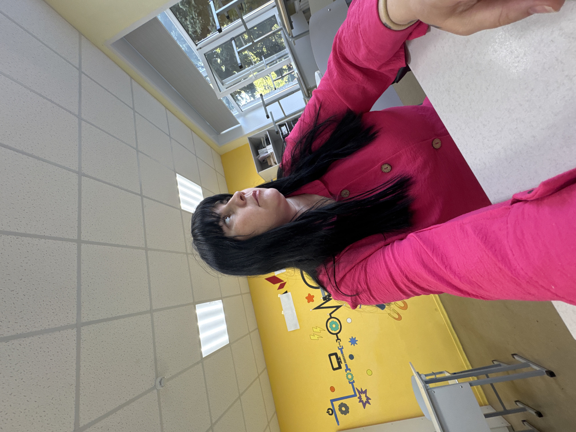 A medium shot presents a woman with black hair and bangs, wearing a bright pink button-down shirt. She is seated at a white table in what appears to be a classroom. The room has white, tiled ceilings and a yellow wall adorned with cartoonish drawings of gears and shapes. A window is visible in the background. The setting is likely Zav'yalovo, Russia.