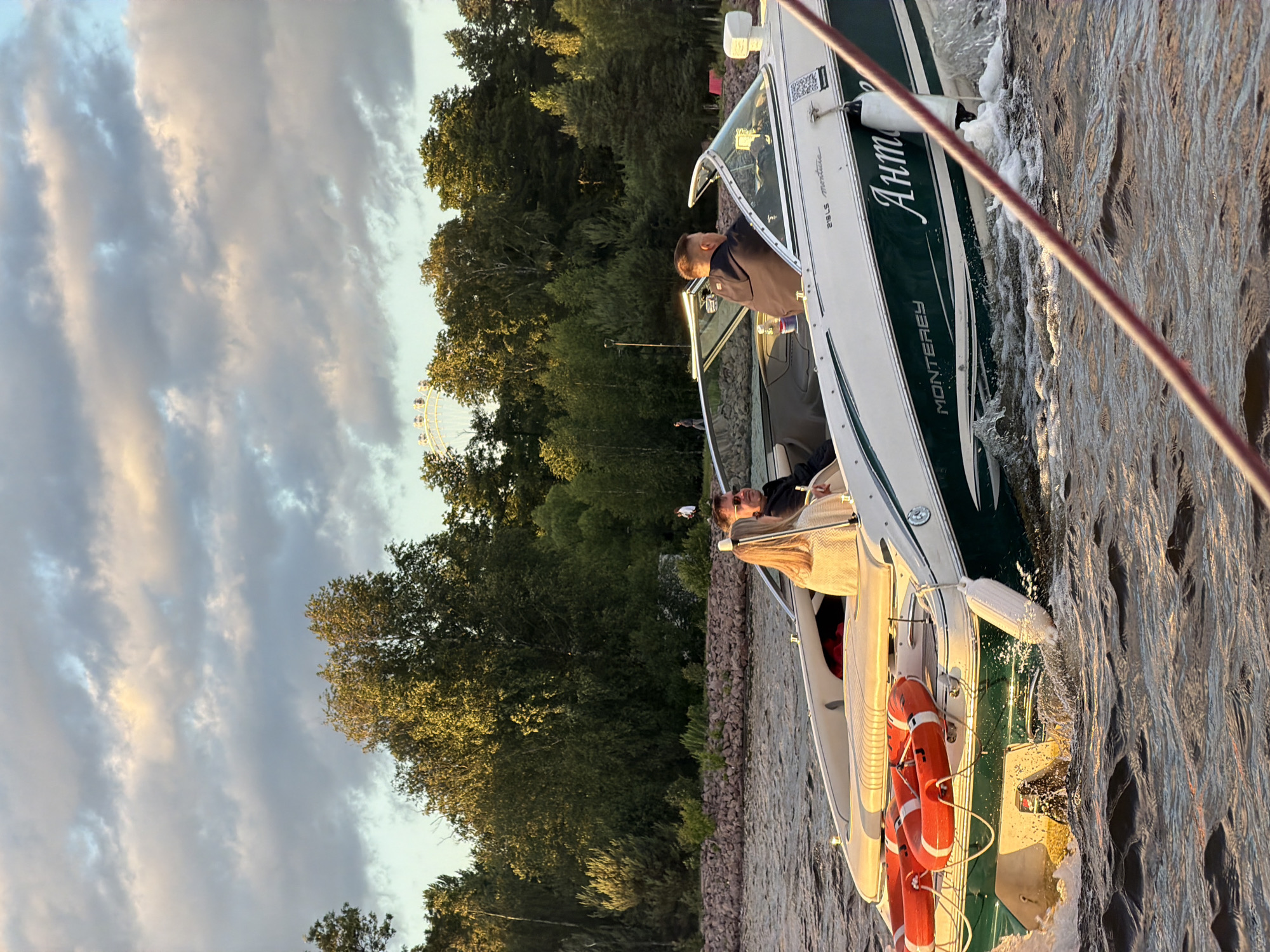 A group of people are on a boat in the waters of Saint Petersburg, Russia. The boat, a green and white Monterey with the name "Антон" written on its side in cursive, is moving through choppy, sunlit water, creating a wake. Three life preservers are attached to the back of the boat. A woman with blonde hair is in the passenger seat, wearing a cream-colored knit top. A man in a dark jacket is driving the boat. Behind them, another person in a dark jacket is visible. In the background, lush green trees line the shore, and a large Ferris wheel is visible in the distance, suggesting the boat is near an amusement park or recreational area within the city. The sky is partly cloudy with soft, warm light filtering through, indicating it may be late afternoon or early evening.