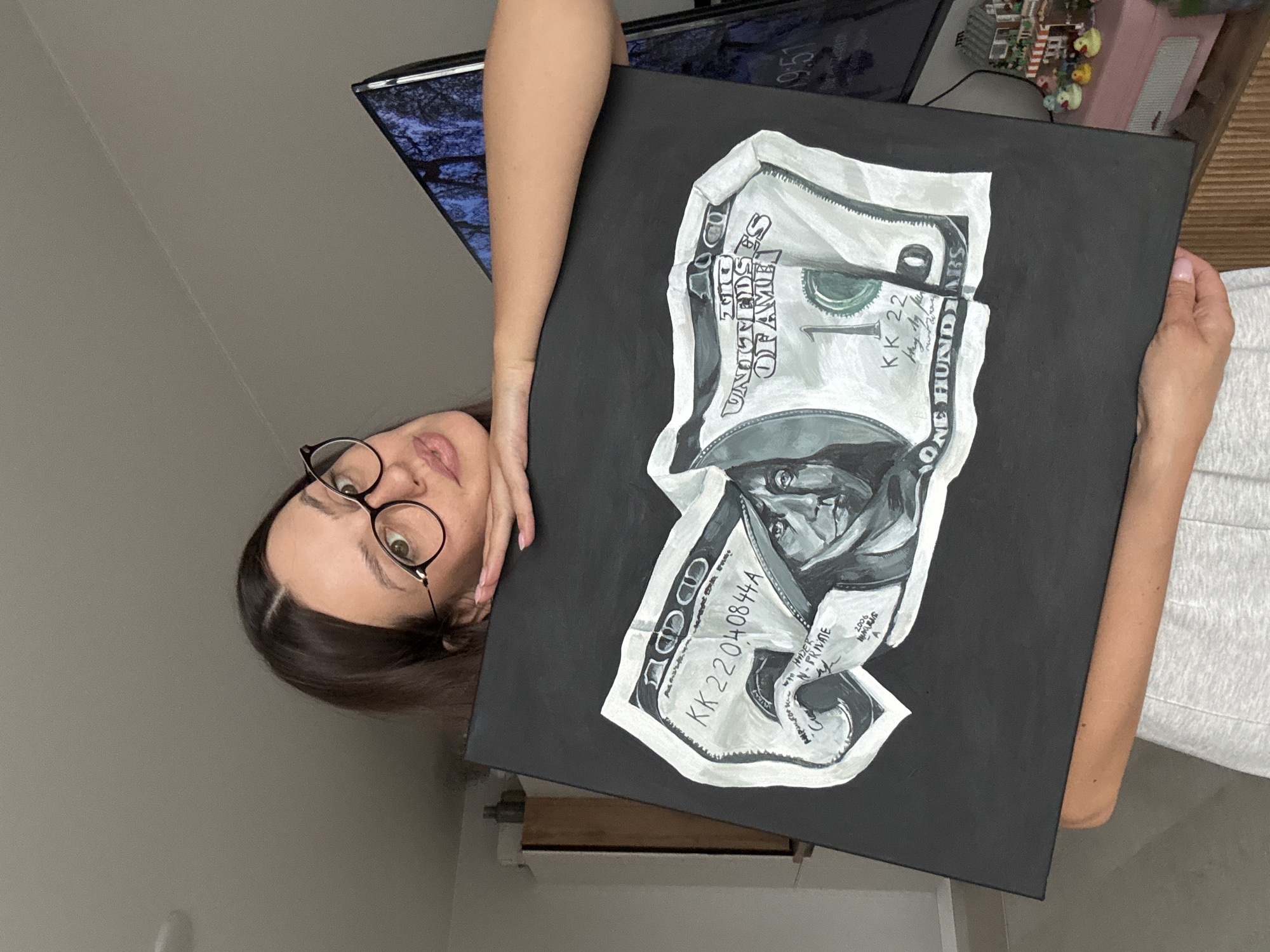 A woman with dark hair, wearing round glasses and a grey t-shirt, holds up a painting of a crumpled US one hundred dollar bill on a black canvas. The painting is rendered in grayscale with hints of green. The woman is leaning her arm on top of the painting, looking at the camera with a slight smile. In the background, a television screen displays a dark, abstract image, and a pink cabinet with decorative miniature houses and ducks is visible. The scene appears to be an indoor setting, possibly a living room or studio, with a wooden cabinet and a wall in the background. There is no discernible information in the image that would indicate a specific location within Krasnoyarsk, Russia.