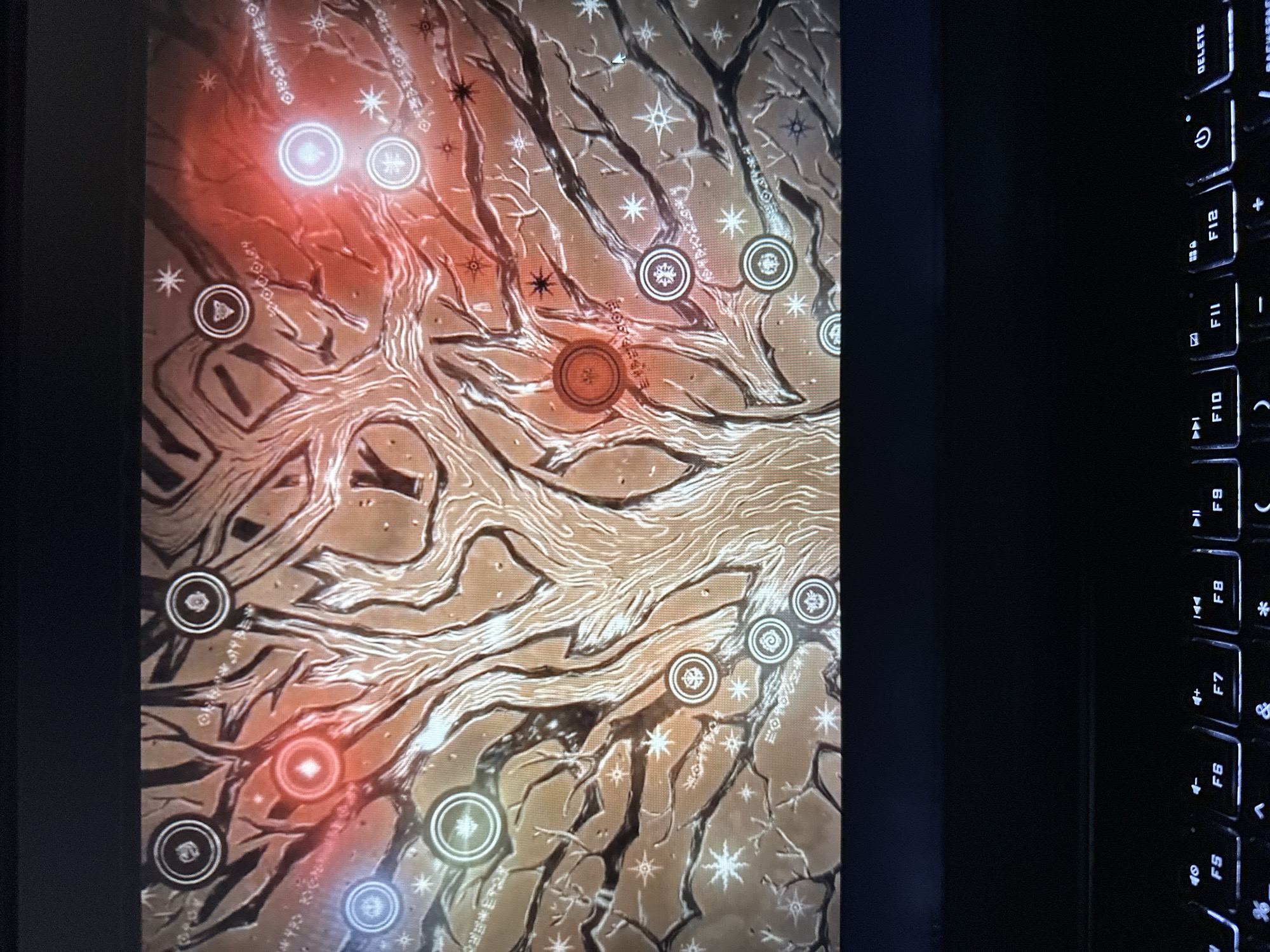 The image shows a close-up of a laptop screen displaying a complex, stylized tree-like diagram. The diagram features interconnected branches with circular icons and glowing orbs placed at various points. The color palette consists of warm tones like orange and red, contrasted with cooler blues and white glows. The background appears to be textured and abstract, possibly representing a mystical or game-related interface. The lower portion of the image shows a dimly lit laptop keyboard. There are no people or recognizable landmarks visible that would indicate a specific location in Vancouver, United States of America.