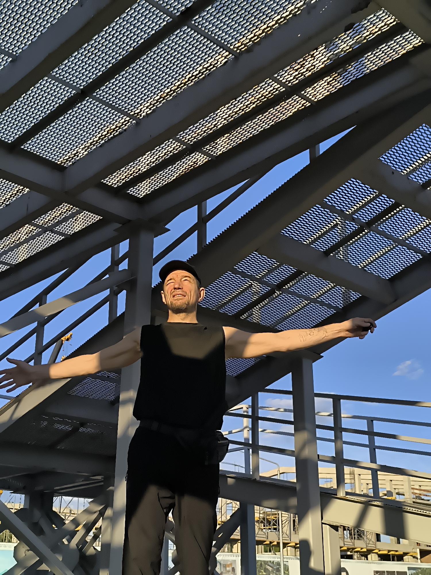 In the city of Qarağandı, Kazakhstan, a man stands on a metal structure under a latticed metal roof. He wears a black sleeveless shirt, black pants, and a black baseball cap. His arms are outstretched to his sides, and he looks up at the sky with his eyes closed and a smile on his face. Sunlight filters through the metal roof, casting dappled shadows on the man and the structure. The sky is a clear blue. The structure appears to be part of an elevated walkway or a similar industrial or public works facility. In the background, there are hints of construction or industrial activity.