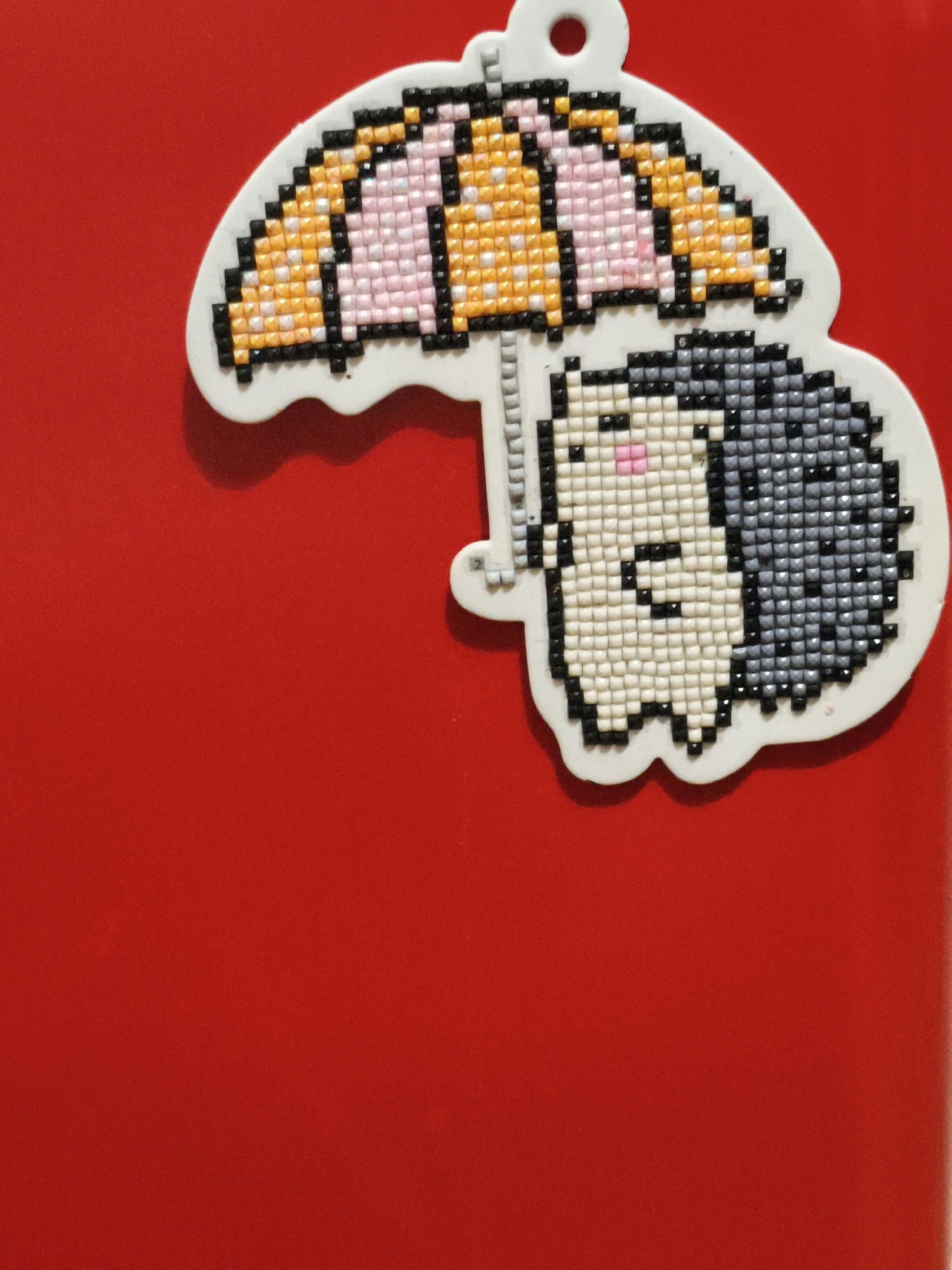 A diamond painting keychain features a cartoon hedgehog holding an umbrella on a red background. The hedgehog is depicted in profile with a gray spiky back, a cream-colored body, and pink cheeks. It holds a yellow and pink striped umbrella above its head. The keychain is shaped like the characters and the umbrella.