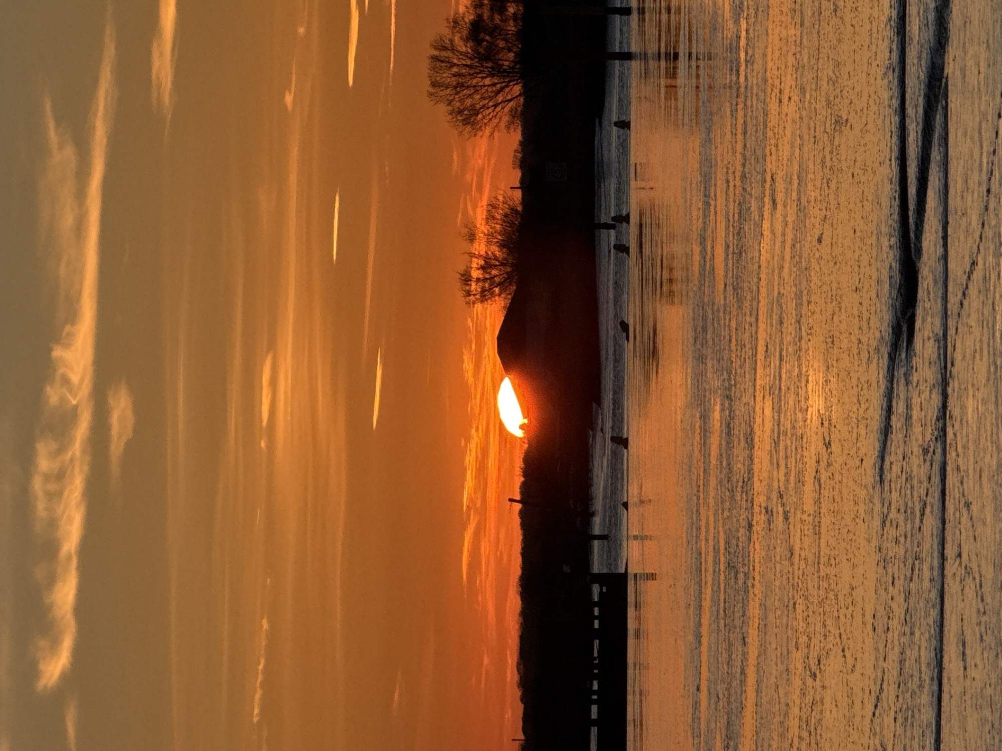 The image captures a sunset over water, possibly a river or harbor, in Great Yarmouth, United Kingdom. The sun is partially obscured by a landmass, casting a warm orange glow on the sky and reflecting on the water surface. Silhouetted trees and posts line the water's edge, creating a stark contrast against the bright sky. Vertical streaks of clouds are visible in the sky, adding texture to the scene. In the foreground, the image shows a textured vertical surface.
