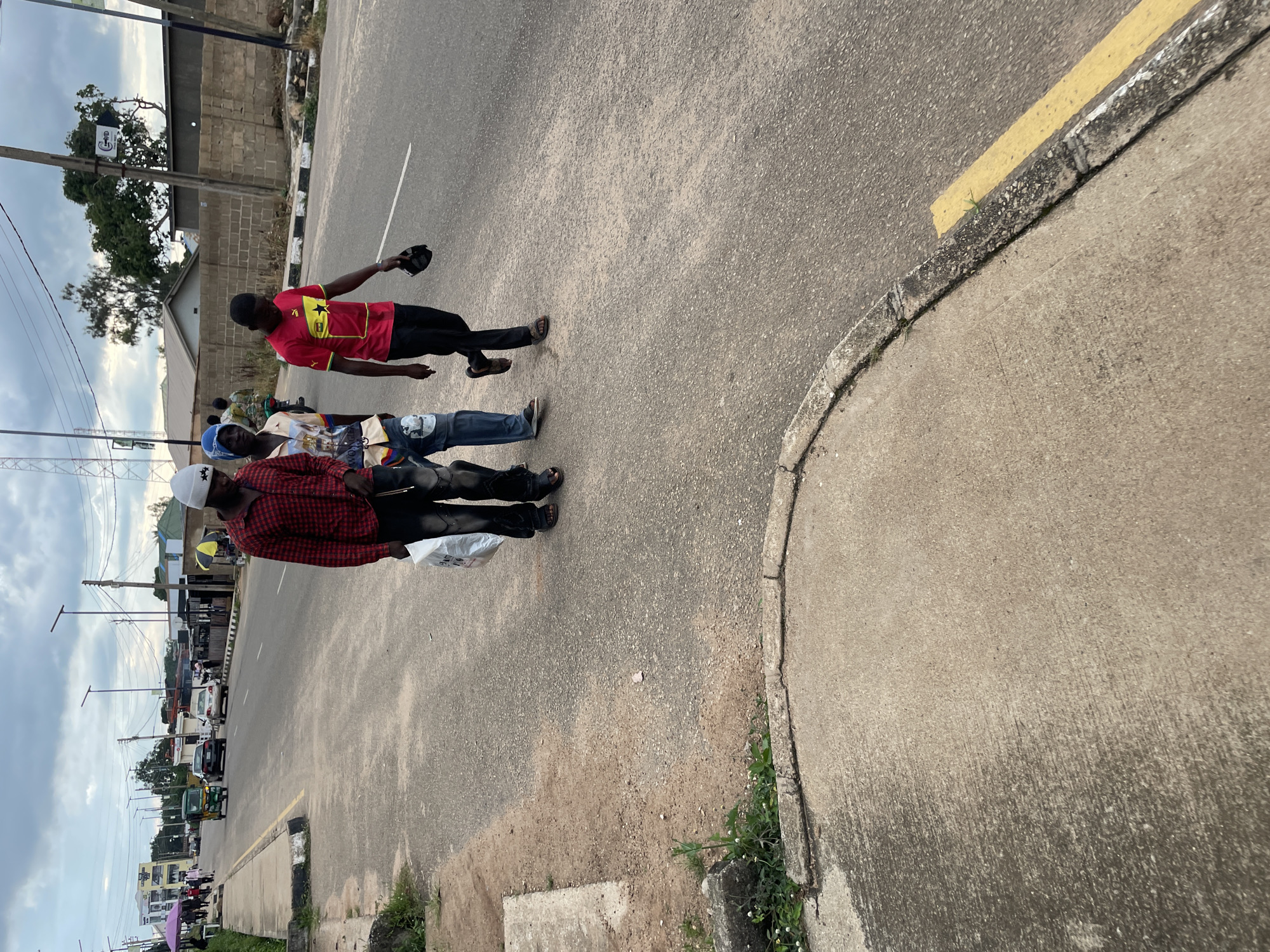 Three young men walk on a paved road in Jos, Nigeria. The man on the left wears a red and black plaid shirt and black jeans, carrying a white plastic bag. The man in the middle wears a light-colored shirt with a graphic print and blue jeans. The man on the right wears a bright red t-shirt with a yellow stripe and a black star, black pants, and holds a black cap. They are walking in the same direction, away from the viewer, on the right side of the road. In the background, vehicles and buildings line the street under an overcast sky. A concrete sidewalk runs along the left side of the road, with some sparse vegetation.