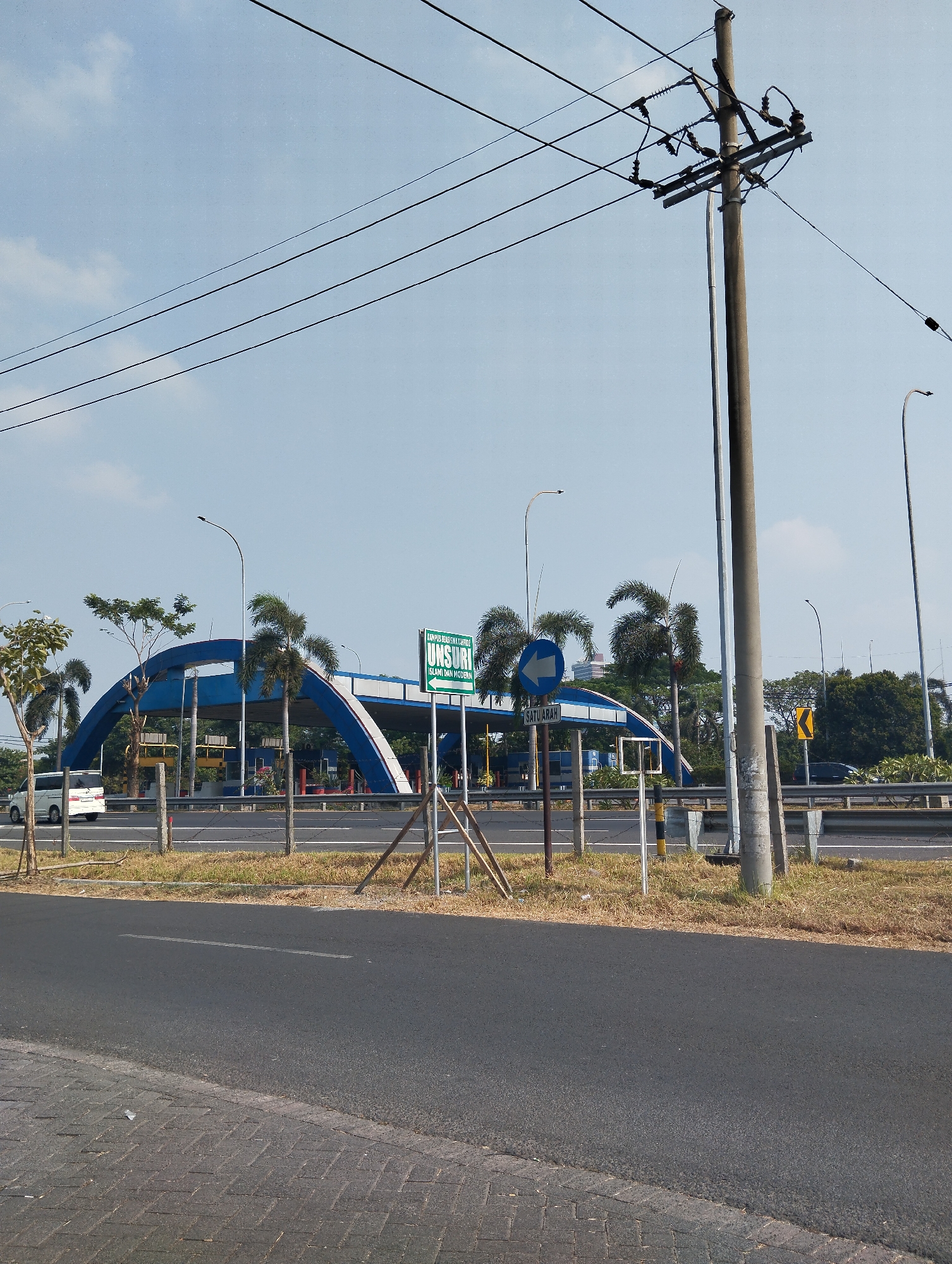 This is a street-level, wide-angle photograph taken outdoors in Surabaya, Indonesia, under a clear blue sky with scattered white clouds. In the foreground, a dark asphalt road occupies the bottom half of the image, with a patterned brick walkway visible in the lower left corner. The road is lined with dry, yellow grass and some scattered debris.
A prominent element is a tall, weathered concrete utility pole on the right side of the frame, supporting multiple black electrical wires that stretch across the sky. To the left, an arched blue structure with white trim spans over what appears to be an entrance or exit to a facility. Several vehicles, including a white van and a dark car, are visible on the road passing under the arch.
In front of the arch, a green directional sign reads "UNIVERSITAS MUHAMMADIYAH SURABAYA (ISLAMI DAN MODEREN)" with an arrow pointing to the left. A blue and white circular sign with a left-pointing arrow and the text "SATU ARAH" (One Way) is also present. Palm trees and other greenery are interspersed around the structure and along the roadside. The scene depicts a well-maintained urban environment with clear signage and infrastructure.