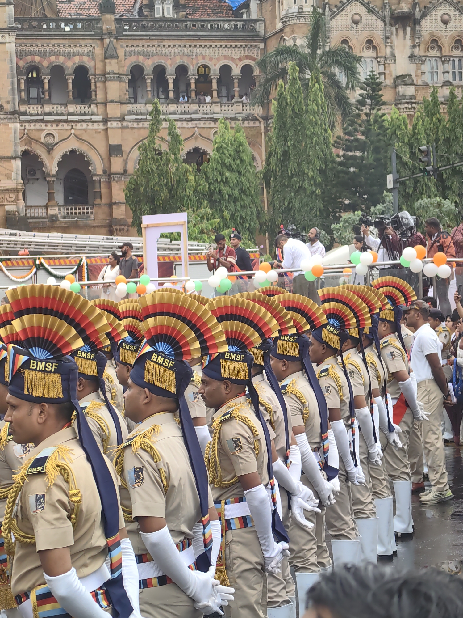 The image depicts a ceremonial event in front of the Chhatrapati Shivaji Maharaj Terminus in Mumbai, India. The foreground is dominated by a line of individuals, appearing to be law enforcement or security personnel, dressed in tan uniforms with distinctive blue and gold regalia on their hats and shoulders. They wear white gloves and stand in formation. Behind them, a crowd of spectators and media personnel are visible, with several individuals holding cameras. Balloons in the colors of the Indian flag (orange, white, and green) are dispersed throughout the scene. The background features the grand architecture of the Chhatrapati Shivaji Maharaj Terminus, a UNESCO World Heritage site, with its ornate stonework and multiple arched windows. The overall atmosphere suggests a public celebration or parade.