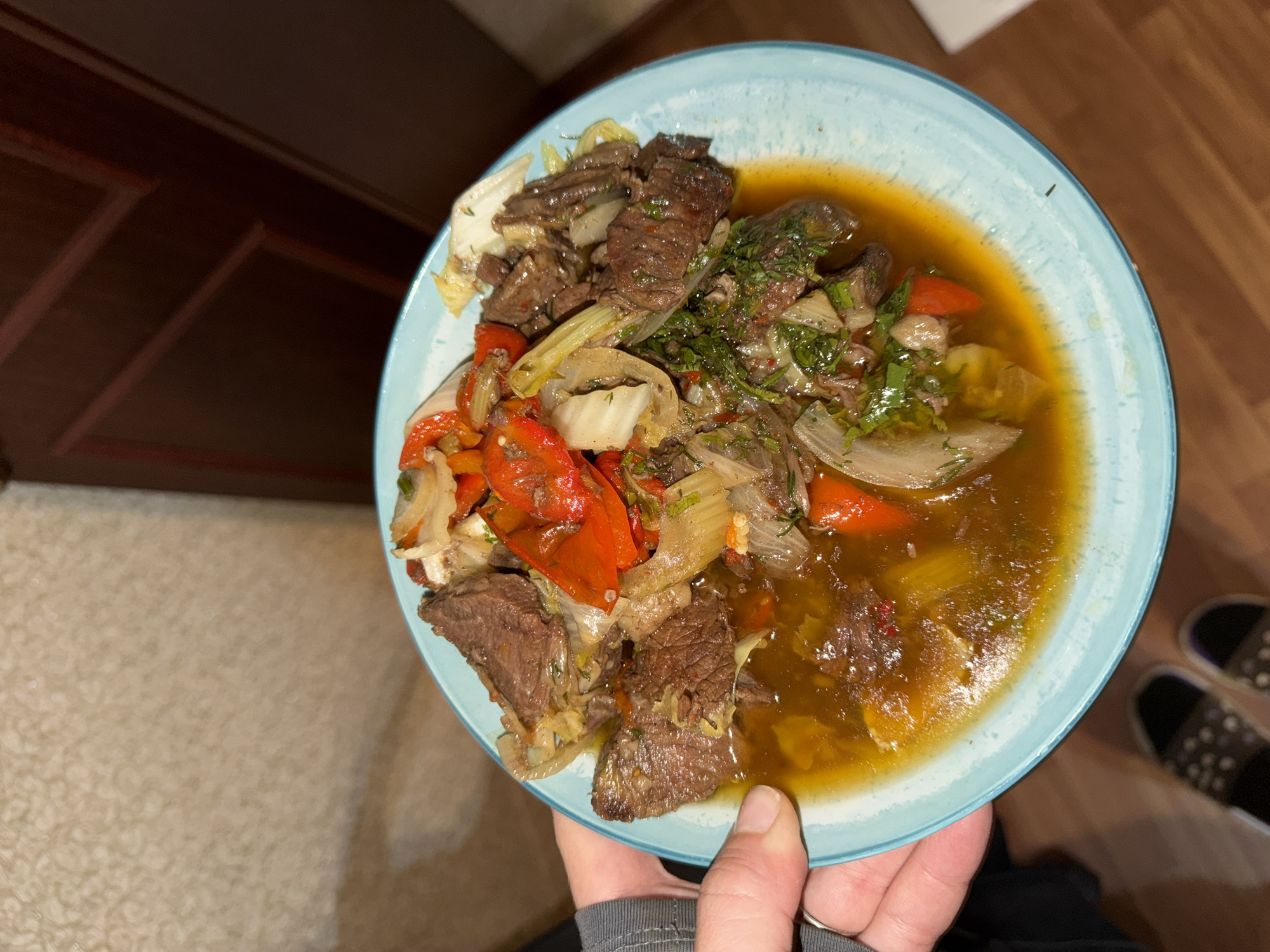 A person is holding a light blue plate filled with a meal consisting of pieces of beef, sautéed vegetables (primarily red bell peppers and onions), and herbs in a brown sauce. The plate is held by a hand with a ring on the finger. The background includes a dark wooden cabinet, patterned carpeting, and a partial view of footwear (possibly slippers). The image does not provide information about the location in Naberezhnye Chelny, Russia.