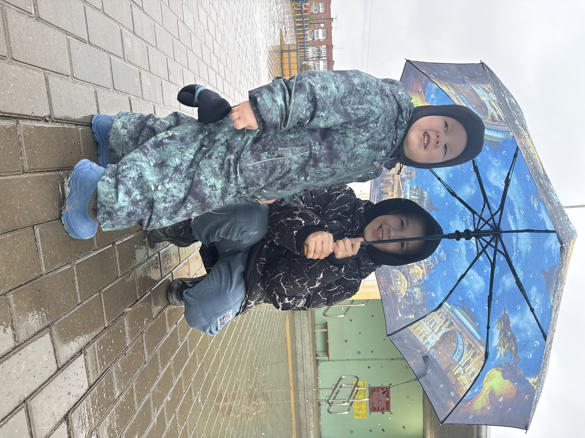 Two young children are positioned on a wet, brick-paved surface, likely a sidewalk or plaza. The child on top wears a patterned green and gray snowsuit with blue boots. This child is leaning on the child below and to the right. The child below wears a black and white patterned jacket, blue jeans, and black shoes. They are crouching while holding a blue umbrella with pictures of Russian architecture. The weather is overcast, with evidence of rain. In the background, there are buildings and some sort of metal barricade with signage. The location is possibly near a building in Zav'yalovo, Russia.
