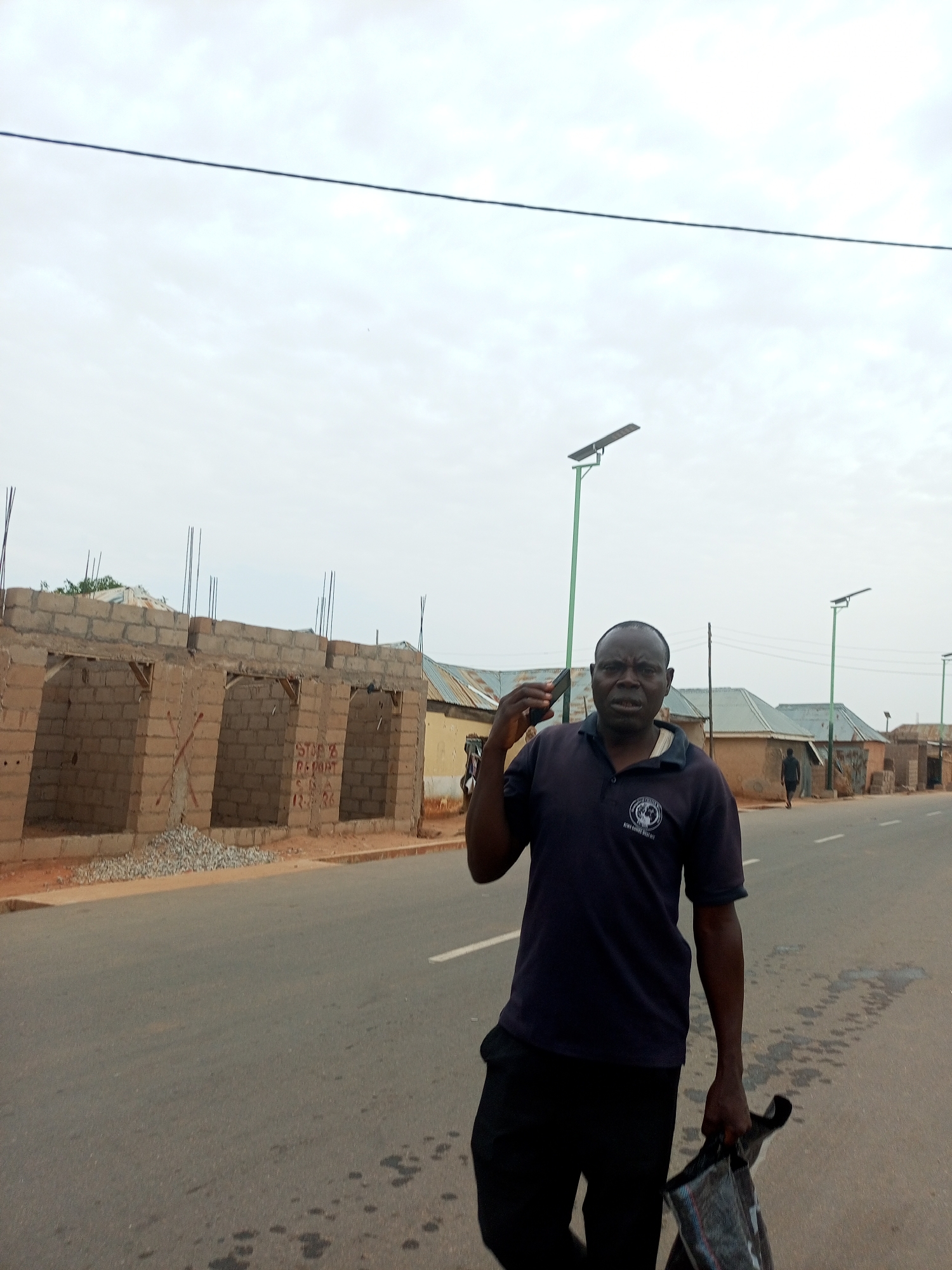 The image shows a man on a road in Gombe, Nigeria. He is wearing a dark polo shirt and dark pants and holding a bag. He appears to be talking on a cell phone. Behind him are unfinished buildings with exposed brick and rebar, along with existing structures typical of a residential or mixed-use area. The road appears to be asphalt with some wear. Utility poles line the road. The sky is overcast.