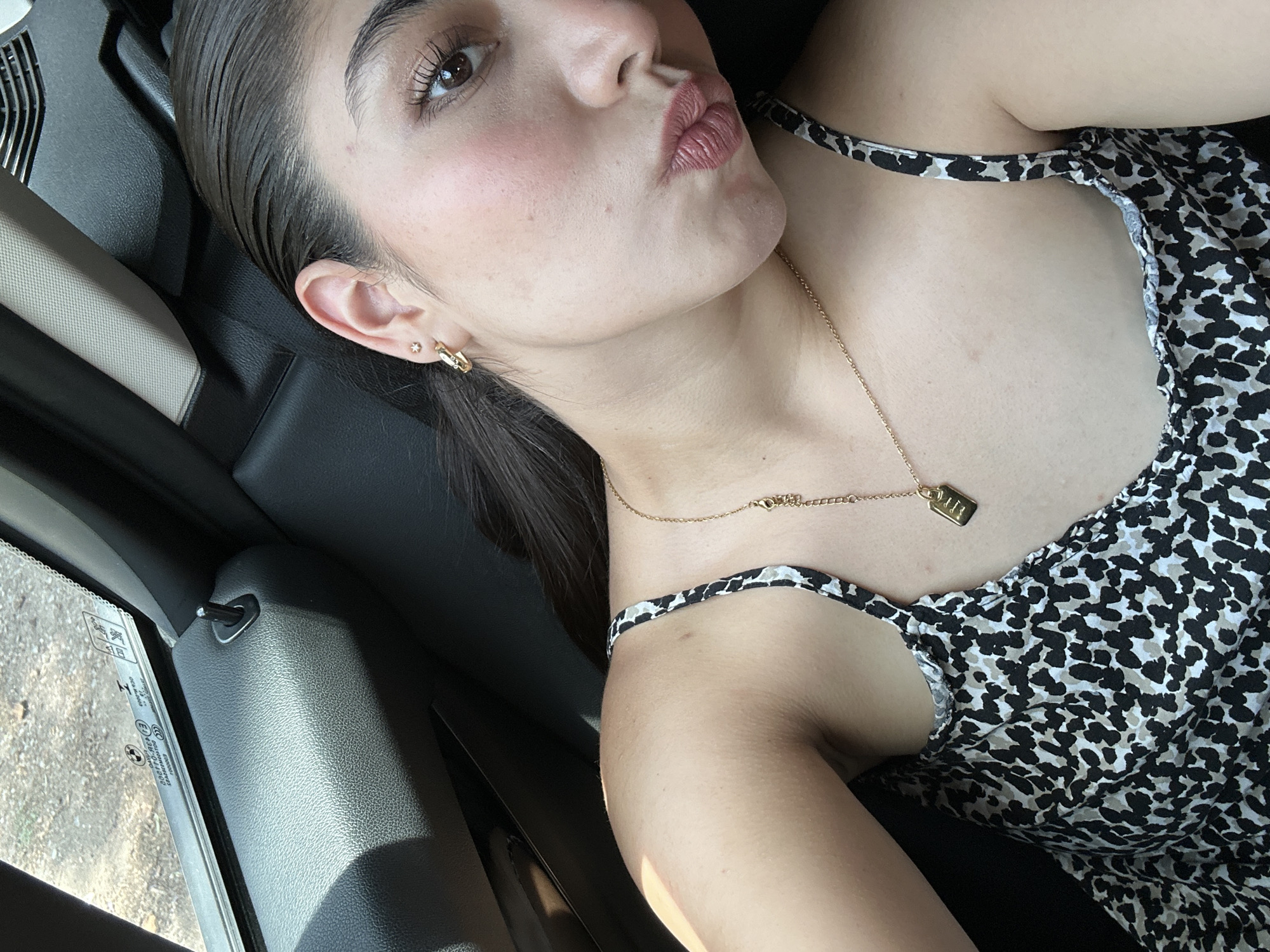 A woman is lying on the black leather seat of a car, looking up at the camera. She is wearing a patterned top and a gold necklace with a small rectangular pendant. Her lips are pursed in a kiss. The interior of the car is visible, including the window and door panel. The location is Unknown, Unknown.