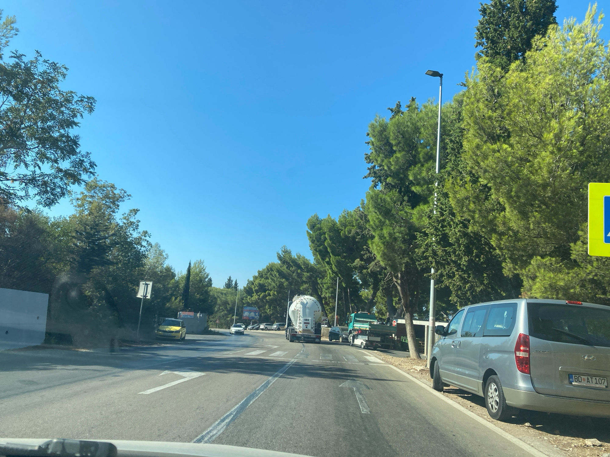 The image depicts a view from inside a vehicle traveling on a road in Sutomore, Montenegro. The road is paved with asphalt and marked with white lines, indicating traffic lanes. On the right side of the road, a silver Hyundai minivan is parked on the shoulder. The minivan has a license plate that reads "BD AT 107". Beyond the minivan, there are trees lining the side of the road.
The road ahead shows multiple lanes with directional arrows painted on the surface. Traffic appears to be moving, with several vehicles visible in the distance. These include a white truck carrying a large cylindrical tank, a green dump truck, and other passenger cars. The environment is characterized by lush green trees, a clear blue sky, and bright sunlight, suggesting a sunny day. There are streetlights and various road signs visible. The overall scene is that of a typical road in a town or city, with civilian vehicles and infrastructure.