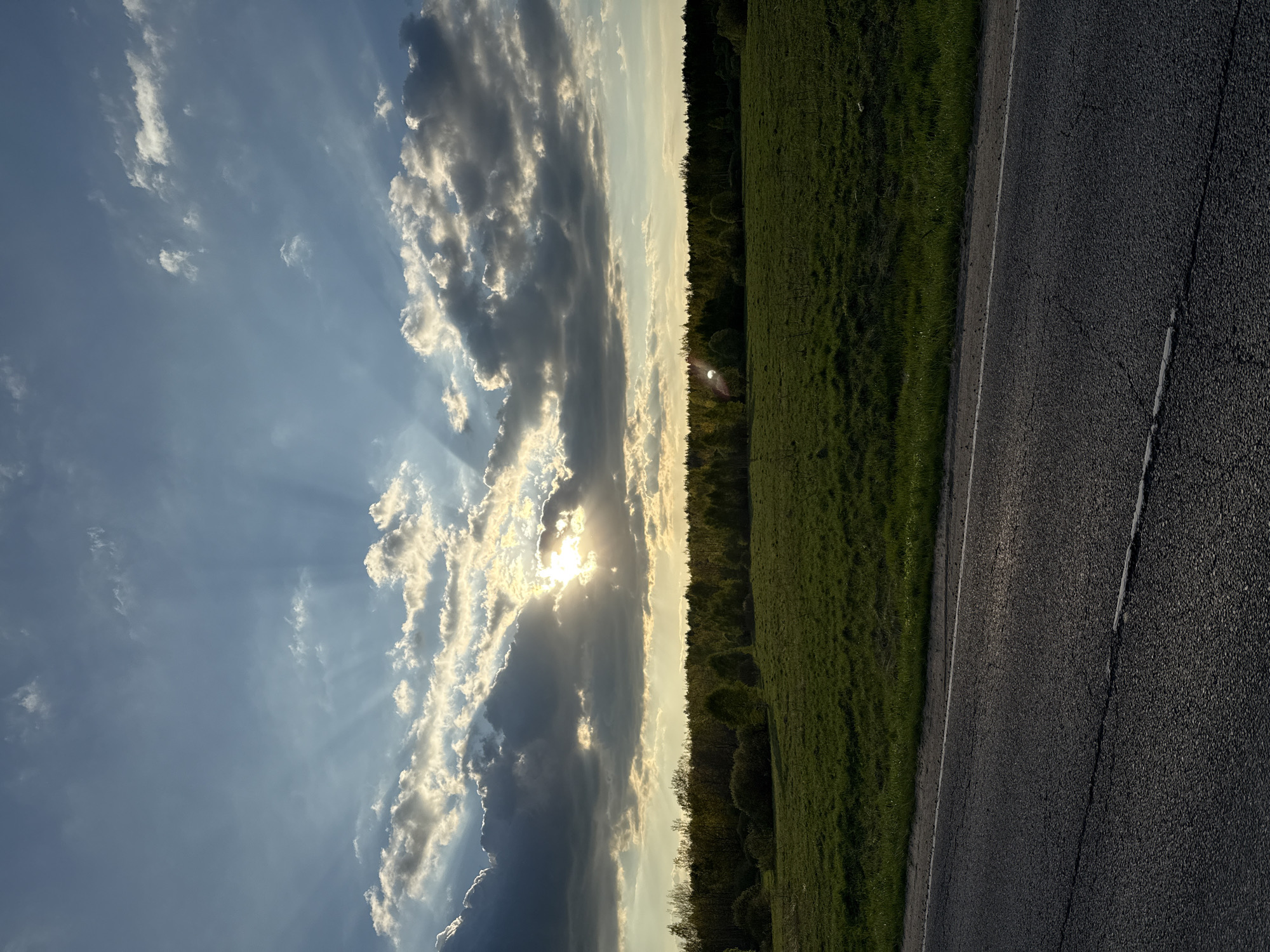 The image depicts a landscape with a view of the sky, a field, a line of trees, and a portion of a road. The sky is filled with clouds illuminated by sunlight, with sun rays visible through gaps in the clouds. A green field extends along the right side of the image, bordering a line of trees that appears to be a forest edge. To the right of the field, a portion of an asphalt road is visible, showing its dark surface and a painted line marking the edge. The composition is taken from a low angle, likely at ground level. There are no visible people. The scene is likely in a rural area with natural light. It is not possible to confirm the location within Rastovtsy, Russia with the information available in the image.