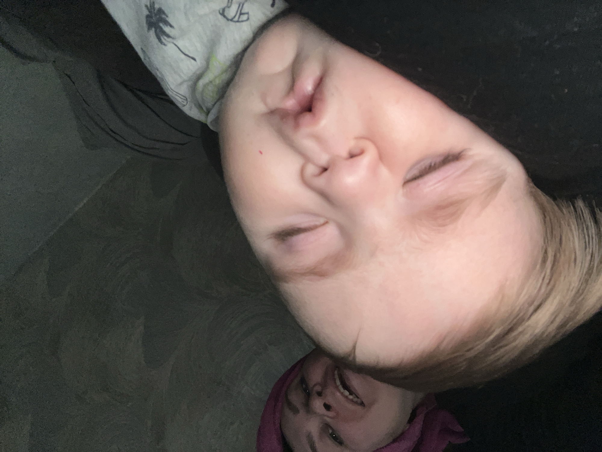 A photograph taken indoors, rotated approximately 90 degrees clockwise, shows a child asleep on a person wearing a purple head covering. The child has blonde hair and is wearing a grey shirt with a palm tree design. The person wearing the head covering is smiling. The background includes what appears to be a grey surface. There is no information to determine if this is in Noviy, Russia.