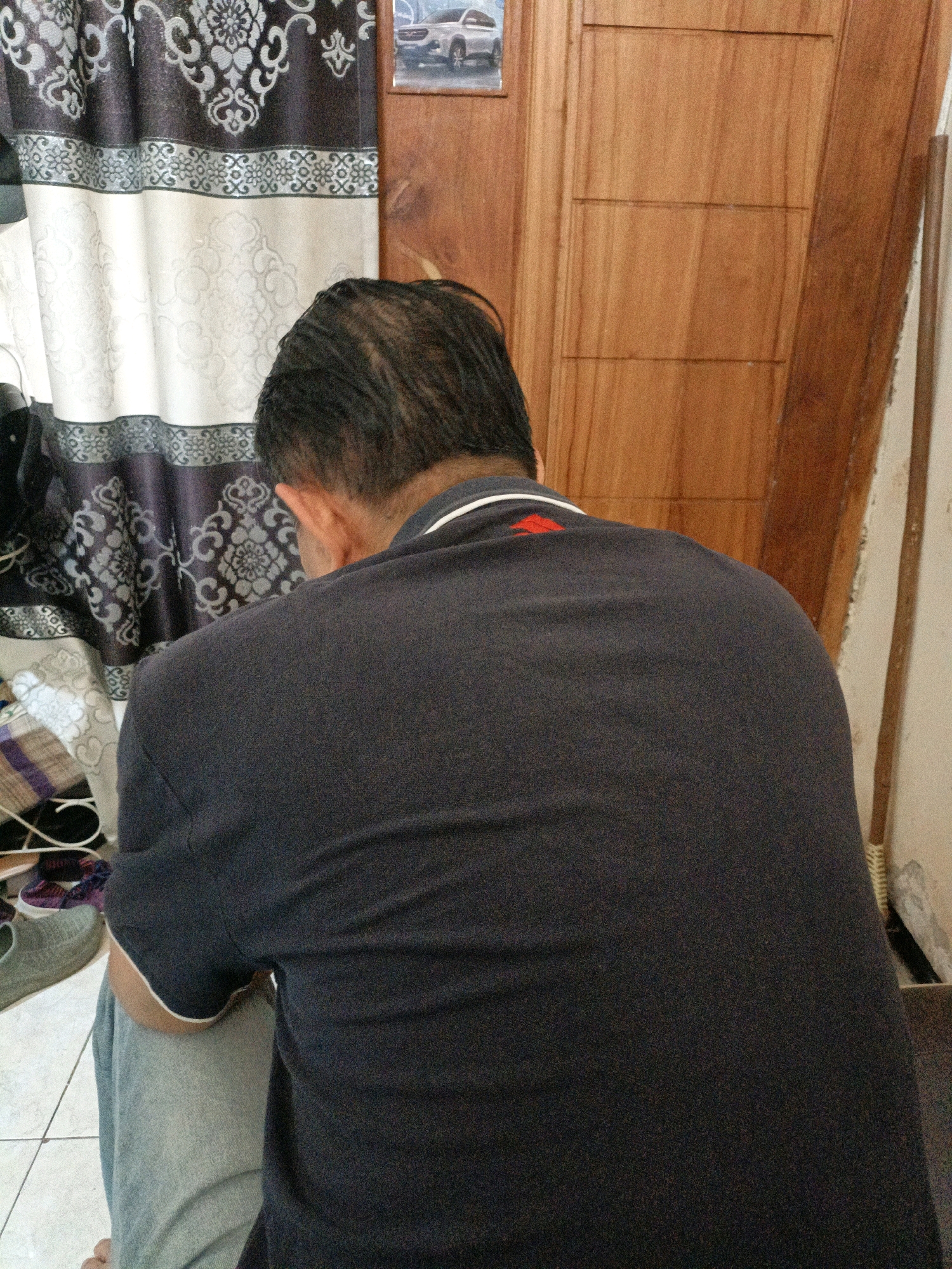 The image depicts an indoor scene, possibly a home in Sidoarjo, Indonesia. The foreground is dominated by the back of a person wearing a dark blue polo shirt with a white collar and a red emblem on the left shoulder. The person's head is bowed, obscuring their face. They are wearing light blue pants. To the left of the person, a patterned curtain with floral motifs in black and white is visible, partially obscuring a collection of shoes and bags on the floor. Behind the person, a wooden door with horizontal paneling occupies the right side of the frame. A framed picture of a white car is affixed to the top left portion of the door. The wall to the right of the door is painted a light color and has a wooden pole leaning against it. The flooring appears to be white tiles.