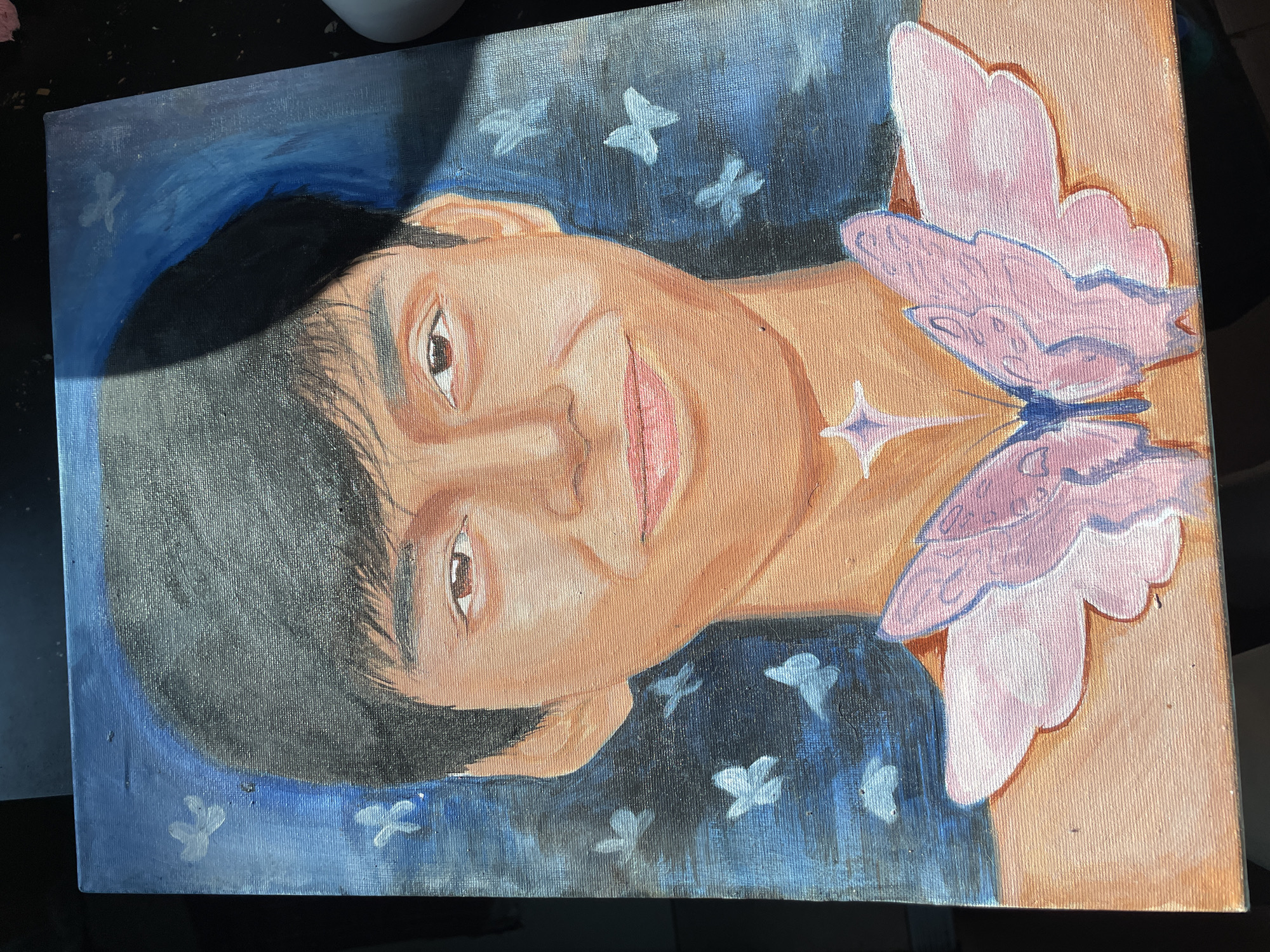 This is an image of an acrylic or oil painting on a canvas with a visible weave texture, located in Davao City, Philippines. The painting depicts a head-and-shoulders portrait of a young individual with short dark hair, brown eyes, and light brown skin, exhibiting a slight smile. The subject's bare shoulders are visible, adorned with a large, stylized pink butterfly motif centered on the chest and a glowing four-pointed star on the neck area. The background of the painting is dark blue, featuring multiple scattered light blue and white butterfly shapes. The physical painting is illuminated by a bright, directional light source from the upper right, casting a shadow on the upper left portion of the canvas, and is resting on a dark, indistinct surface.