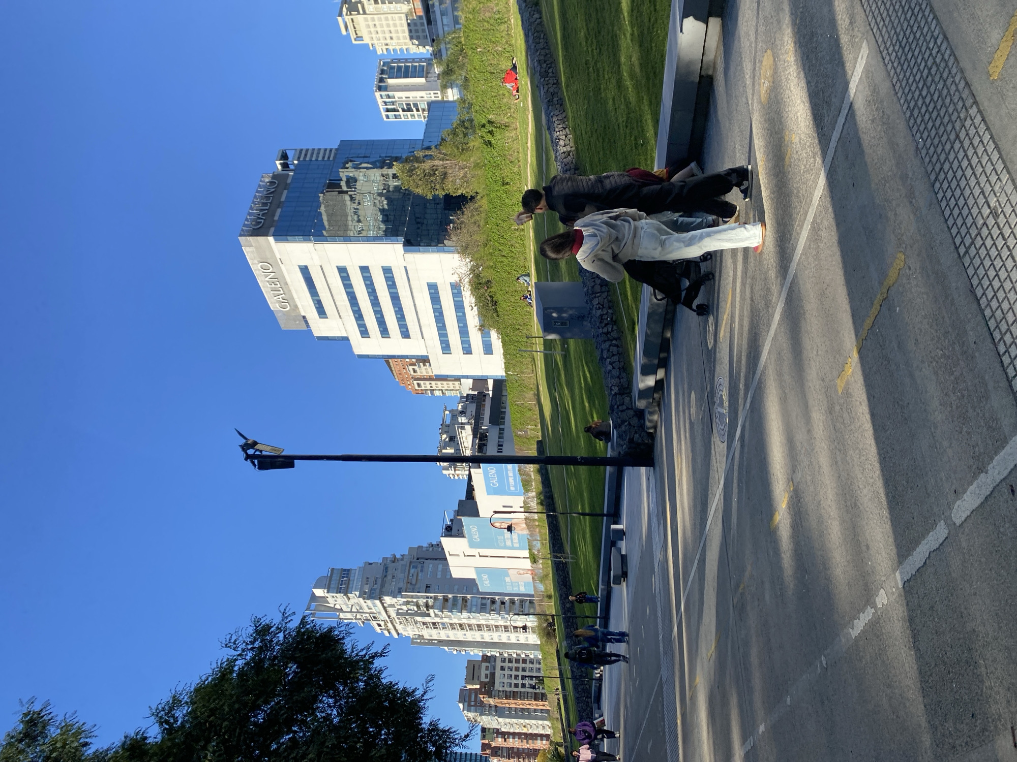 The image depicts a street scene in Buenos Aires, Argentina, identifiable by the "GALENO" signage on a tall, modern building. The scene shows a paved walkway with white and yellow painted lines, alongside a grassy area bordered by rocks. Several people are present: two individuals are leaning against the rocks, one in a light-colored jacket and the other in dark clothing, seemingly engaged with their phones. Other people are walking along the pavement in the distance. A black lamp post stands prominently in the foreground. The background showcases numerous high-rise buildings under a clear blue sky.