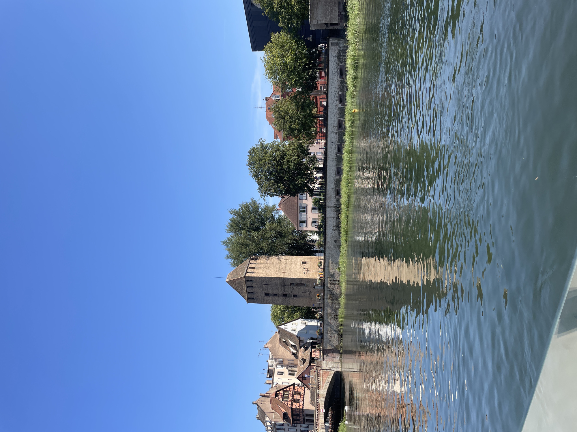 The image captures a picturesque view of the Petite France district in Strasbourg, France, on a clear, sunny day. The foreground is dominated by a tranquil body of water, likely a canal or river, with ripples reflecting the surrounding architecture and greenery. Along the bank, a stone embankment is visible, topped with lush green grass and reeds.
Dominating the mid-ground is a historical stone tower, characterized by its square base, weathered appearance, and a pyramidal roof. Adjacent to the tower are several charming buildings with characteristic Alsatian architecture, featuring steeply pitched roofs, exposed timber framing on some, and warm, earthy color palettes. A stone bridge arches gracefully over the water to the left, connecting the riverbanks and providing a pathway.
Trees with full green canopies are interspersed along the embankment, casting shadows and adding to the scenic beauty. The sky above is a vibrant, cloudless blue, providing a serene backdrop. The reflection of the buildings and the tower is clearly visible in the water, creating a mirror image that enhances the depth and tranquility of the scene. No people are clearly discernible in the frame. The overall impression is one of historical charm and natural beauty.