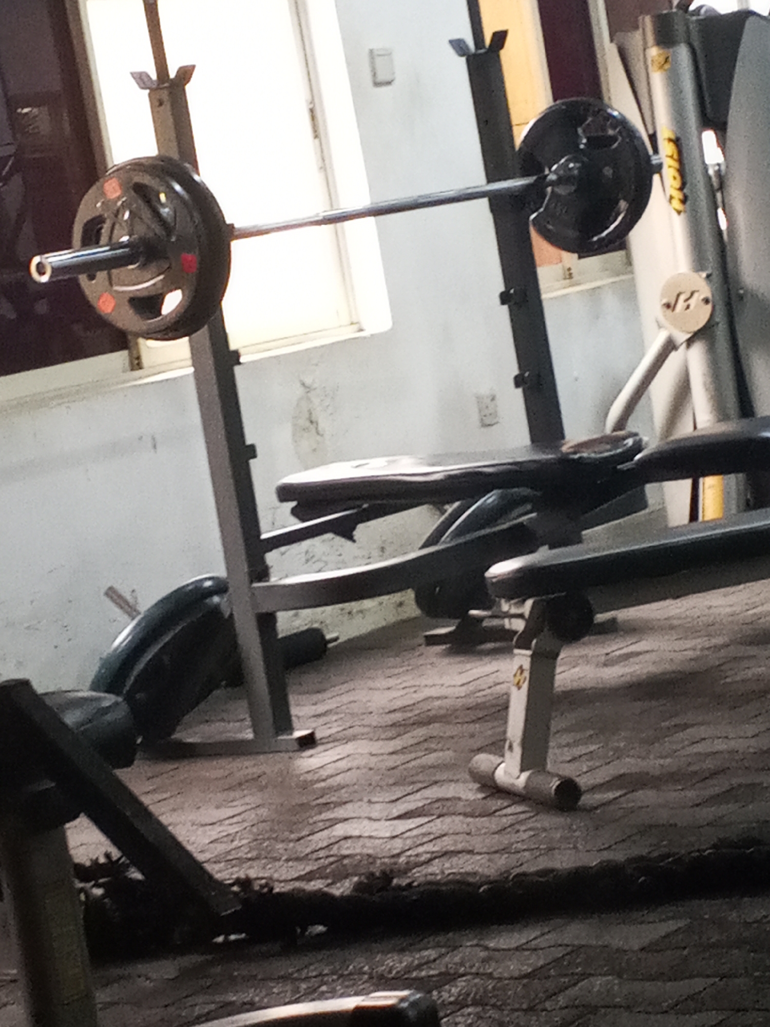 This image depicts a gym setting in Abuja, Nigeria, likely indoors. The primary objects in focus are weightlifting equipment, specifically a barbell loaded with weight plates resting on a rack, and a weight bench. The barbell is positioned horizontally, supported by the rack which appears to be part of a bench press station. One weight plate on the left side of the barbell is partially visible, showing orange markings. The weight bench, a dark-colored piece of equipment, is positioned in front of the rack, suggesting it's set up for bench presses.
In the background, a large window allows natural light to enter the space, creating a bright, almost blown-out effect. The walls appear to be painted a light color and show some signs of wear or staining, contributing to a lived-in gym aesthetic. To the right of the bench press setup, another piece of gym equipment is partially visible, possibly a leg press or similar machine, identifiable by its metallic structure and gray color. The floor is covered with a textured surface, possibly rubberized mats or a specialized gym flooring, which has a patterned appearance.
On the left side of the image, another section of gym equipment is visible, including what appears to be a leg curl or extension machine, dark green in color and angled towards the floor. The overall lighting suggests it could be daytime, though the indoor setting makes it difficult to pinpoint the exact time. There are no people visible in the frame, indicating the image was taken in an empty gym or during a time when it's not in use. The overall atmosphere is functional and utilitarian, typical of a fitness facility. No legible text is clearly discernible in the image, aside from potential brand markings on the equipment which are too blurry to read.
