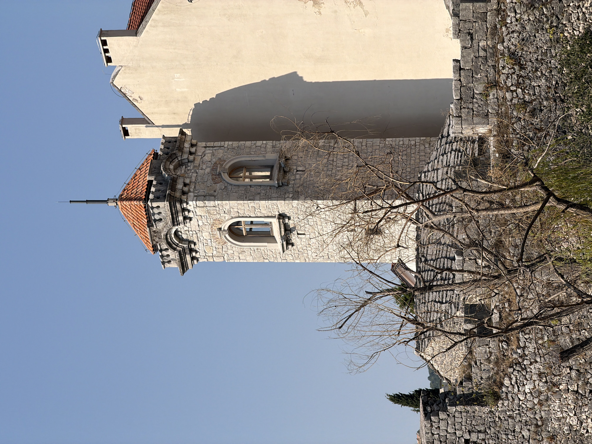 The image shows a stone tower with a red-tiled roof and arched windows, part of a larger stone building, with a beige wall casting a large shadow to its right. In the foreground, there is a stone wall and the bare branches of a tree. The location is the city of Trebinje, Bosnia and Herzegovina, specifically the Arslanagić Bridge area, with the tower likely being part of the Old Town fortifications or a related historical structure. The sky is clear and blue.