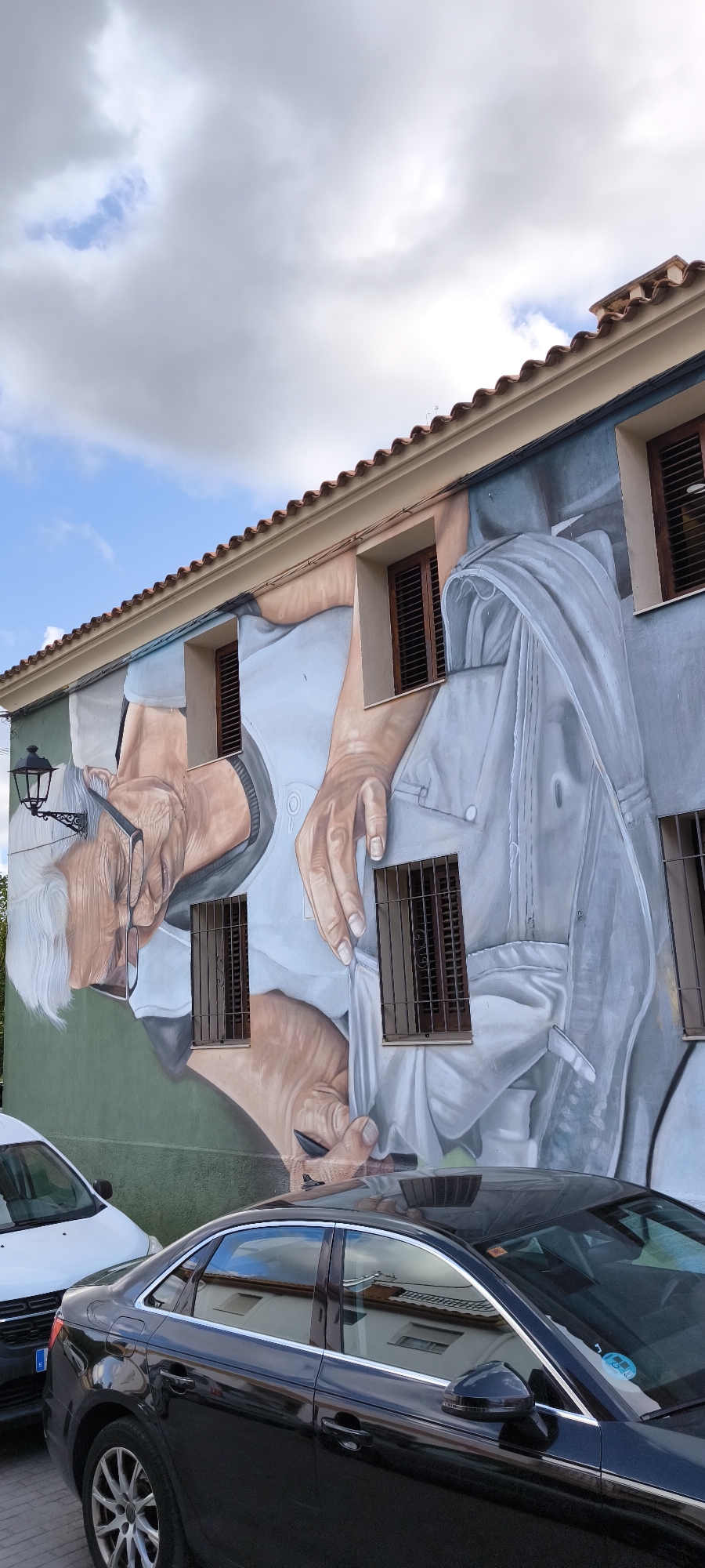 This is a photograph of a building in Fanzara, Spain, featuring a large mural painted on its facade. The mural depicts a close-up of an elderly person's face and hands, rendered in a realistic style with muted colors. The person is wearing glasses and appears to be concentrating on something. The mural is integrated with the building's architecture, with windows passing through the painted image. The building has a tiled roof and the surrounding environment includes a street with cars parked in front of the mural. The sky above is partly cloudy. The mural occupies a significant portion of the visible wall.