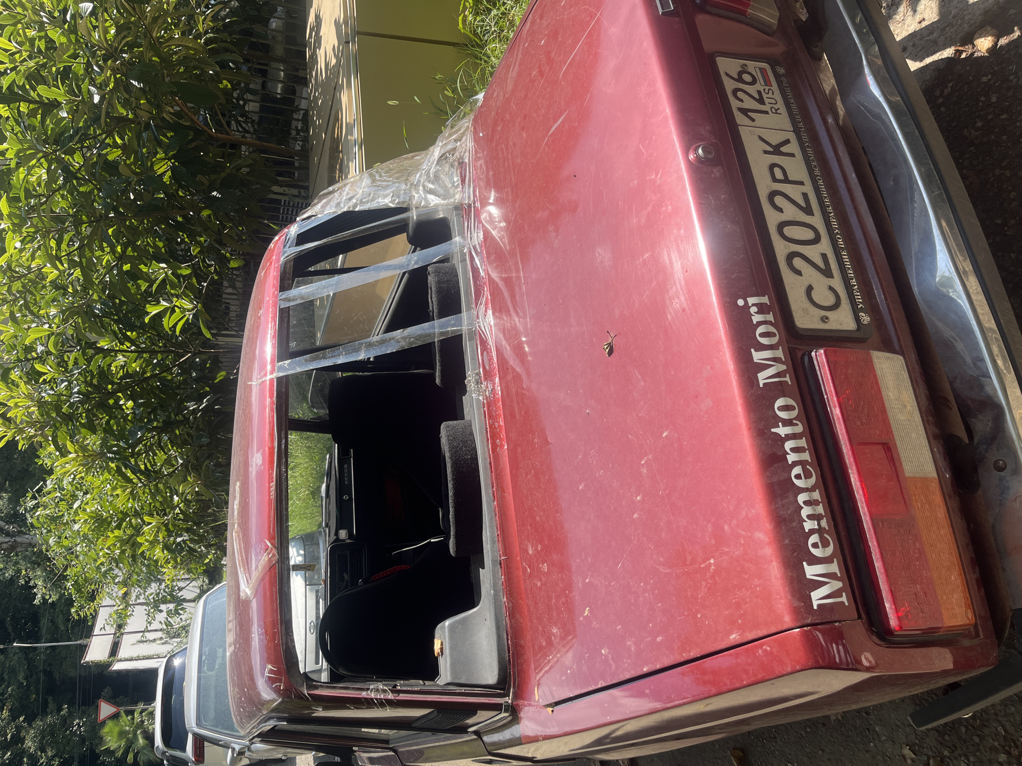 The image shows the rear of a red Lada car parked on a street in Sochi, Russia. The car has a broken rear window covered with clear plastic sheeting. A sticker on the trunk reads "Memento Mori". The license plate is visible and reads "C202PK 126 RUS". In the background, there are lush green trees and a white fence. Two other cars are parked behind the Lada. The overall scene depicts an outdoor environment with a focus on the damaged vehicle.