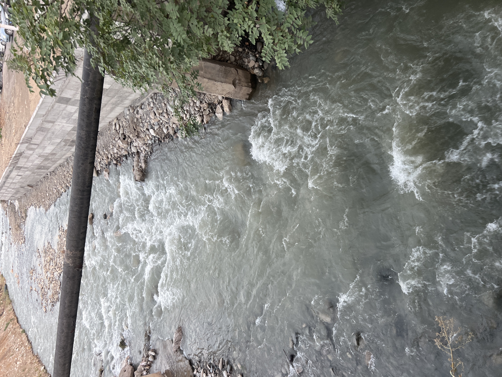 This is an aerial view of the Ak-Bura River flowing through Osh City, Kyrgyzstan. The river is a turbulent, greyish-brown color with white foam and ripples indicating a fast current. Along the right bank, a concrete wall has been constructed, part of which appears to be a retaining wall for a road or building. A large, dark, cylindrical pipe runs horizontally across the middle of the image, supported by a concrete pillar on the right bank and disappearing off the left edge. Vegetation, including leafy green trees and some dry brush, is visible on the right bank. The riverbed is rocky, with visible stones and pebbles. In the distance, on the right side of the image, parked cars and what appears to be a construction site are visible beyond the retaining wall.