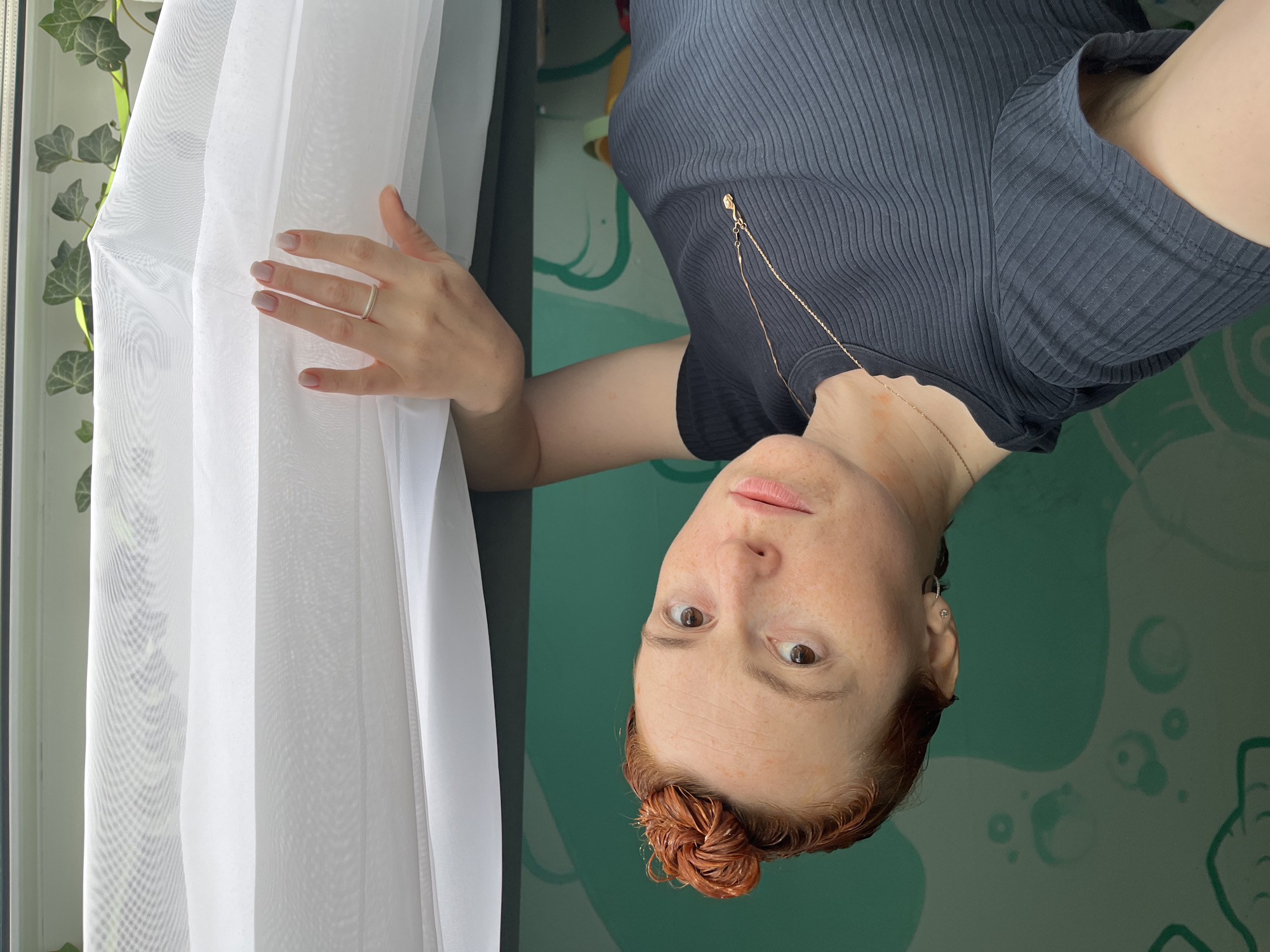The image shows a young woman with reddish hair tied in a bun on top of her head. She is wearing a dark gray ribbed t-shirt and a delicate gold necklace with a pendant. She has freckles on her face and shoulders. Her left arm is bent, and her hand is resting on a sheer white curtain. She is wearing a silver ring on her left ring finger. The background appears to be an indoor setting with a green wall that has a decorative pattern. To the right of the woman, the sheer curtain is illuminated by natural light, revealing a glimpse of green ivy leaves outside the window. The image does not contain any elements that would allow for the specific location in the city of Chaikovsky, Russia to be identified.
