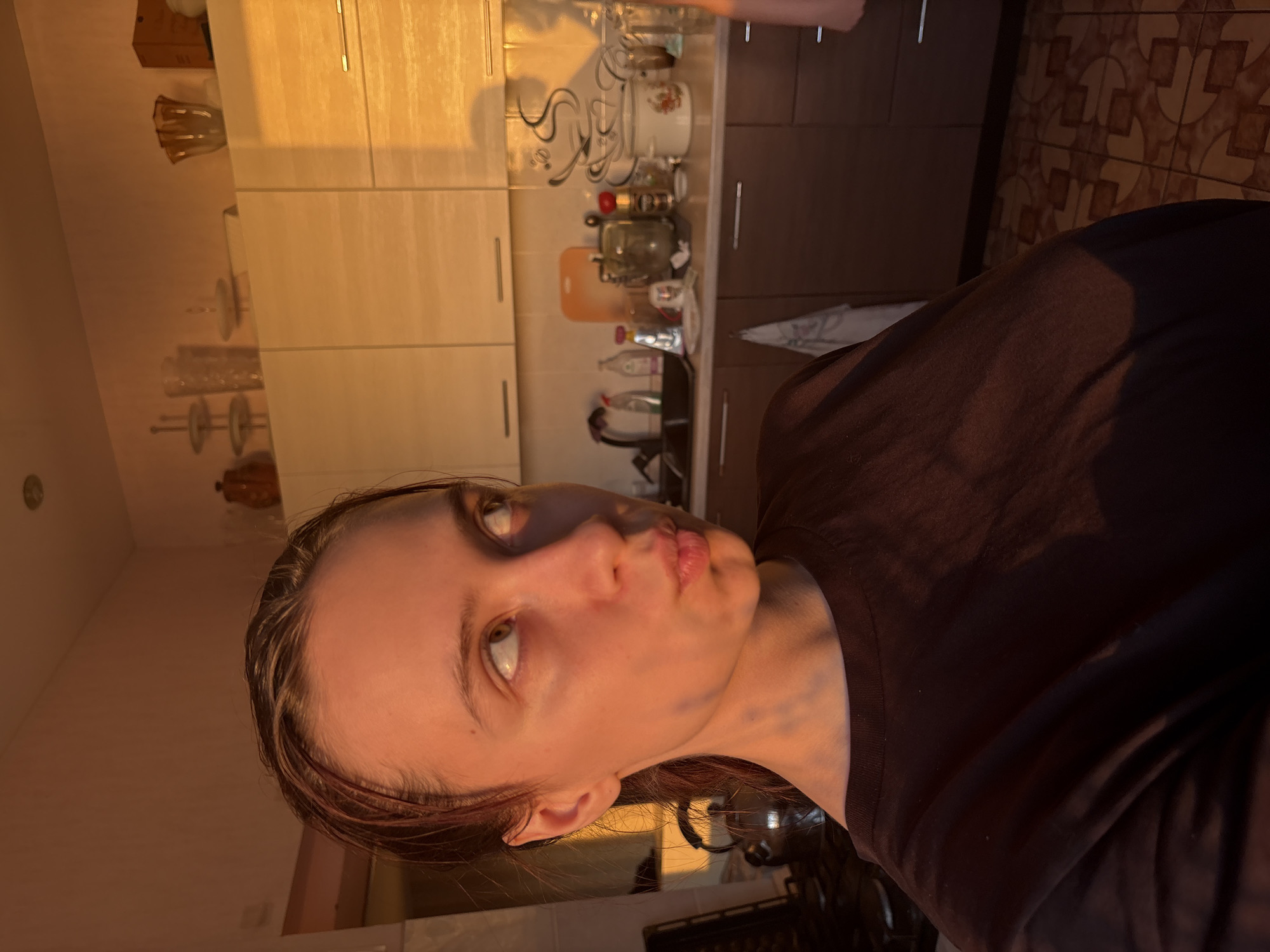 The image is a selfie of a fair-skinned person with reddish-brown hair, wearing a dark t-shirt. The person is facing the camera with a slight upward tilt to their face and a pouting expression. The background is a kitchen, featuring light-colored cabinets, some containing glassware and other kitchen items. A countertop is visible with various bottles and containers. The lighting suggests either early morning or late afternoon, casting a warm, golden hue over the scene. A tiled backsplash and a dark cabinet are also visible in the background. The image was taken in Прикамский, Russia.