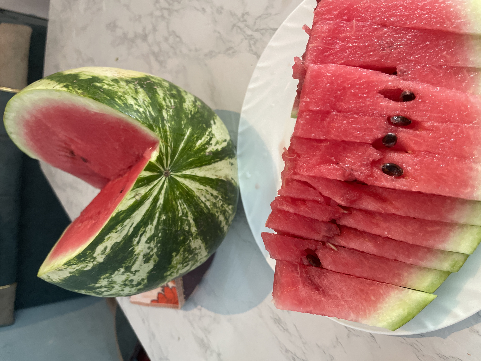 The image displays a close-up view of a watermelon. A large, whole watermelon with a distinctive striped green rind and a slice cut out is positioned in the upper portion of the frame. The exposed flesh of the watermelon is a vibrant pink color. In the foreground, a white plate holds several slices of watermelon, neatly arranged. The slices show the pink flesh and green rind. The scene is set on a marble-patterned surface, likely a table. The background is blurred, showing hints of a teal-colored object and a lighter-colored wall. There is no indication of the city of Bujum, Kyrgyzstan, or any people in the image.