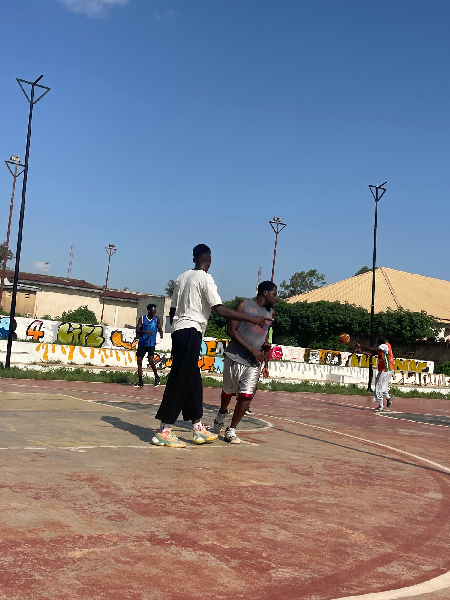 The image depicts an outdoor basketball court in Jos, Nigeria. Several young men are engaged in a basketball game on a reddish-brown asphalt court with white boundary lines. The court is surrounded by a low white wall adorned with colorful graffiti. In the background, there are buildings with light-colored walls and a prominent yellow, sloped roof. The sky is a clear, bright blue with a few wispy clouds. Two tall streetlights, each with a triangular fixture, stand on either side of the court. The players are casually dressed in athletic wear. One player in the foreground, wearing a white t-shirt and black pants, is facing away from the camera, seemingly participating in the game. Another player in a gray tank top and silver shorts is looking towards the ball, which is held by a player wearing an orange and white jersey. The overall atmosphere suggests a lively and active environment.