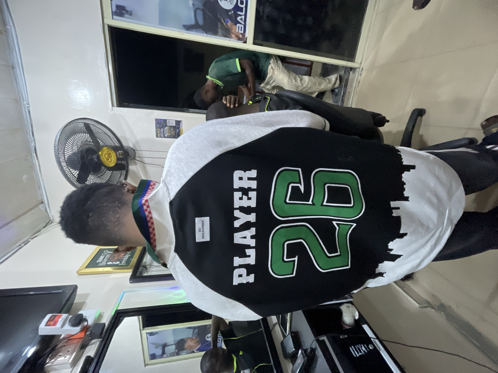 A person is getting a haircut in a barbershop. The barber is giving the customer a cut in front of a window. The customer is wearing a black and white jersey with the word "PLAYER" and the number "26" on the back. A fan is visible in the background. The barber is visible in the reflection on the mirror. The exact location in the city of Ado, Nigeria, cannot be determined from the image.