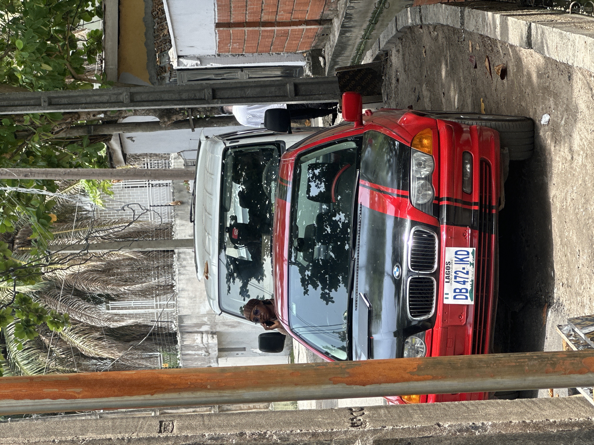 The image captures a street scene in Lagos, Nigeria, featuring a red and black BMW parked adjacent to a white van. A person is visible inside the van, appearing to be on a mobile phone. The car's license plate is clearly visible, displaying "LAGOS" and the code "DB 472-IKD." The background includes various structures, foliage, and a rusty metal railing in the foreground. The scene is illuminated by bright sunlight.