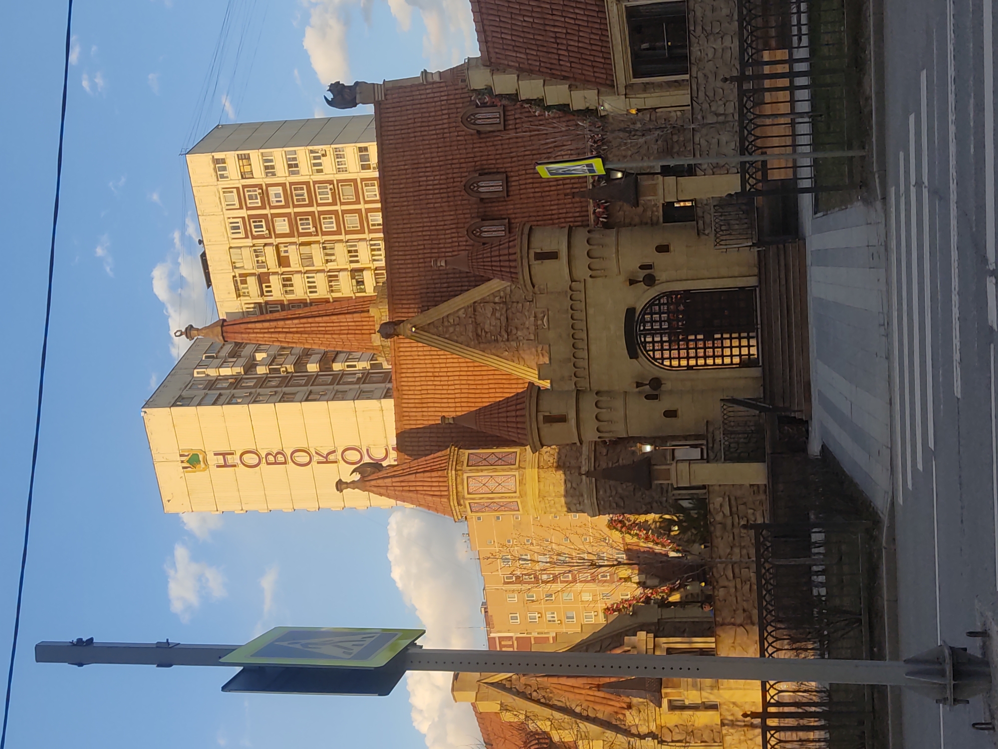 Here's a detailed description of the image:
The image shows a street scene, likely in Moscow, Russia, given the Cyrillic lettering on a building. The scene is slightly tilted.
**Buildings and Architecture:**
* A building resembling a small, stylized castle or tower with turrets and reddish-brown tiled roofing is prominent. This structure has decorative details like arched windows and stonework.
* Behind the "castle" are taller, modern apartment buildings. The buildings are off-white and have a grid pattern of windows. One building has the word "Новоком" (Novokom) vertically displayed in Cyrillic.
* There is a building adjacent to the castle building with similar red-brown roofing.
**Street Elements:**
* A pedestrian crosswalk is visible in the foreground.
* A pedestrian crossing sign (green and yellow diamond shape) is mounted on a pole on the left.
* A decorative metal fence separates the castle structure from the street.
**Environment:**
* The sky is blue with scattered white clouds, suggesting a fair weather day.
* Trees (possibly bare, indicating late fall or winter) are near the buildings.
**Objects:**
* Utility wires stretch across the sky in the top-left.
**In summary:** The image depicts a street in Moscow, Russia, featuring a fanciful castle-like building juxtaposed against modern apartment blocks. A pedestrian crossing and associated signage are visible, indicating a street scene. The lighting suggests it's either early morning or late afternoon.