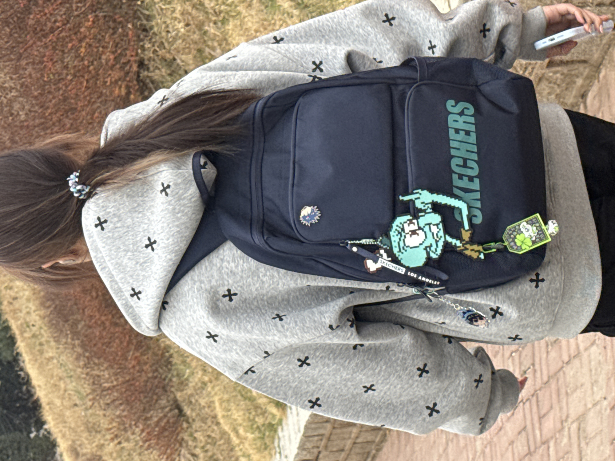 From a high-angle perspective, the image captures a portion of a person situated outdoors, possibly in Zhongtun, China. The person is wearing a grey hoodie adorned with black cross patterns, and a dark blue Skechers backpack. The backpack features the brand name "SKECHERS" in green lettering and is decorated with pixelated cartoon character charms. The person's partially visible hair is tied back with a colorful scrunchie. In one hand, the person holds a mobile phone. The background includes some foliage and a brick-paved surface.