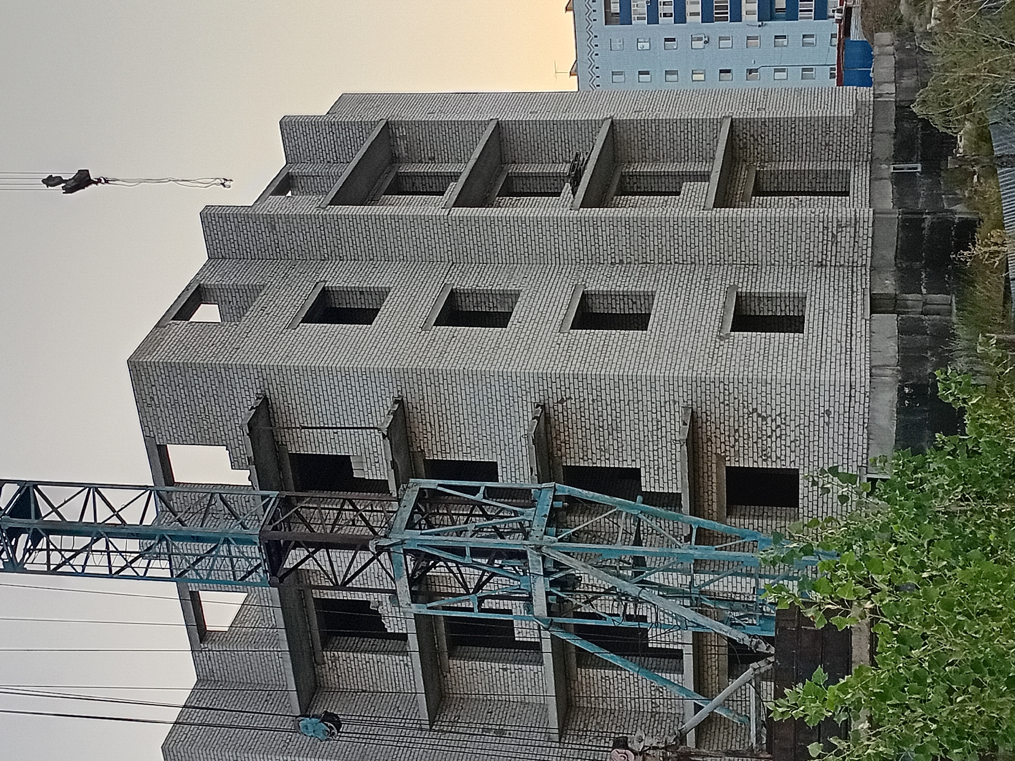 The image shows a multi-story brick building under construction in Semey, Kazakhstan. A blue construction crane is positioned next to the building, with its lattice structure and lifting mechanisms visible. The building itself appears to be made of light-colored bricks and has several floors with open window frames and concrete floor slabs. In the background, a pale blue apartment building with visible windows and air conditioning units can be seen. Green foliage and some dry grass are in the foreground, suggesting an outdoor environment. The sky is a pale, overcast color, indicating a possible dusk or overcast daytime scene.