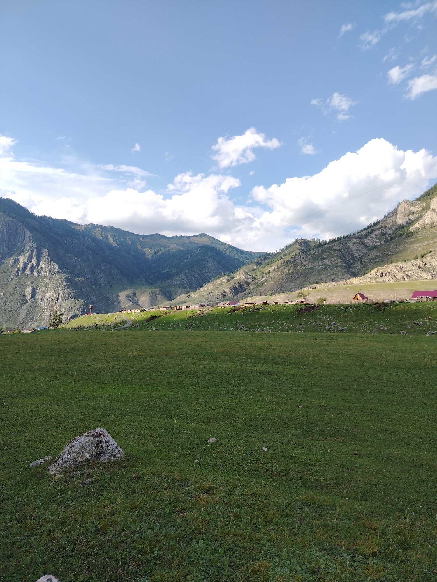 A wide-angle photograph captures a rural mountainous landscape under a partially cloudy blue sky. In the foreground, a vast expanse of vibrant green grass dominates, with a large, grey, moss-covered boulder situated in the lower left quadrant. Beyond the grassy field, a village is nestled on a gentle slope leading up to towering, rugged mountains. The village consists of numerous small, wooden structures with some having red roofs, arranged in a line along the hillside. A dirt road winds through the settlement. The mountains themselves are steep and rocky, with some areas covered in sparse vegetation and trees. The sunlight casts shadows, highlighting the contours of the terrain. The scene suggests a remote, natural setting. This image appears to be taken in Iodro, Russia, judging by the context.