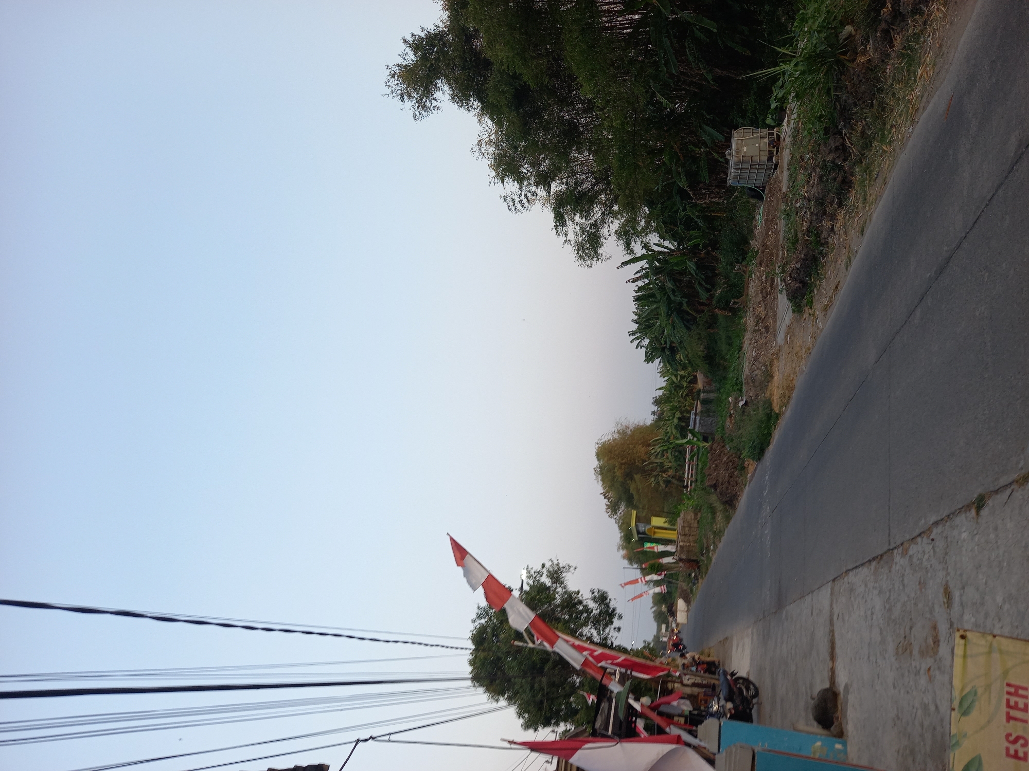 A straight, paved road runs through a rural area in Kebon Agung, Indonesia. The road is bordered on the left by a concrete sidewalk and a dirt shoulder, and on the right by a wooded area. Indonesian flags, red and white, are displayed along the left side of the road. Several trees, including banana trees and bamboo, line the right side of the road. A motorcycle is parked on the sidewalk on the left. A large, industrial-looking tank is situated on the right side of the road. The sky is clear and pale blue, suggesting either early morning or late afternoon. The overall impression is of a quiet, semi-urban or rural setting.