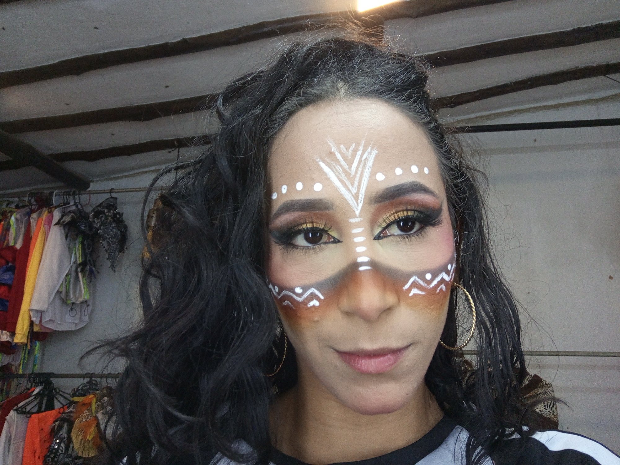 A close-up, eye-level indoor portrait features a woman with elaborate face paint, centered in the frame. Her long, dark, wavy hair frames her face and shoulders. Her face is adorned with white patterns, including an upward-pointing chevron design on her forehead surrounded by white dots, and wavy lines on her cheeks. These white markings are applied over brown and orange shading on her upper cheeks, nose, and around her eyes, creating a tribal or ceremonial appearance. Her eyes are enhanced with dramatic makeup, featuring golden eyeshadow, dark eyeliner, and long, full eyelashes. She has pink blush on her upper cheekbones and softly pink, slightly parted lips. She wears large golden hoop earrings and a top that appears to be black with white stripes or panels.
The background reveals an indoor setting, likely a dressing room or shop, with various garments hanging on racks visible on the left and partially on the right. The ceiling above consists of light-colored panels interspersed with dark wooden beams. A bright light source is visible in the upper right corner. The woman's elaborate makeup and the surrounding context suggest she is prepared for or participating in a performance, cultural event, or celebration in Porlamar, Venezuela. Her expression is neutral to slightly contemplative, looking directly at the viewer. There is no visible text.
