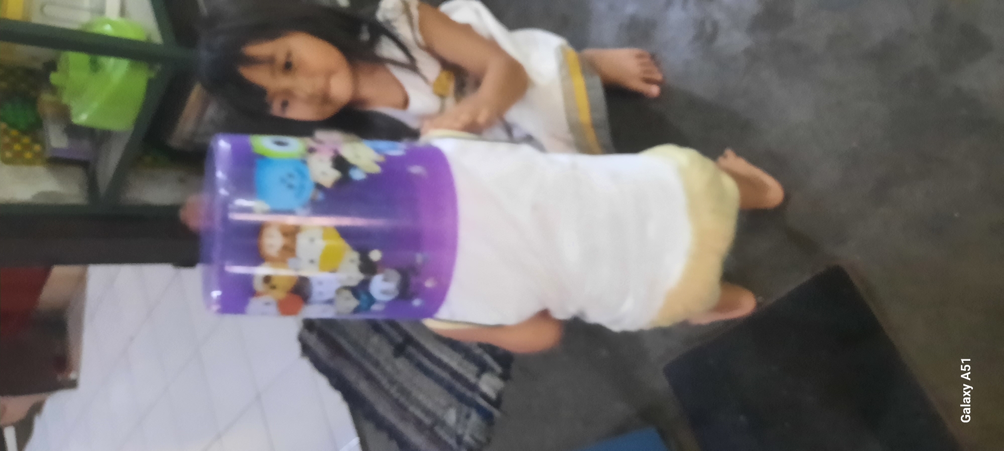 In Jatitengah, Indonesia, an outdoor scene shows two young children in a tiled room. A child with a white shirt and yellow shorts is wearing a large purple plastic container over their head, obscuring their face. The container is decorated with cartoon characters. To the right, another child with dark hair and a white dress with grey and yellow trim looks at the child with the container. The floor is made of grey tiles, and a dark rectangular object is visible in the lower left corner. A striped rug is present in the background to the left. The lighting suggests it is daytime.