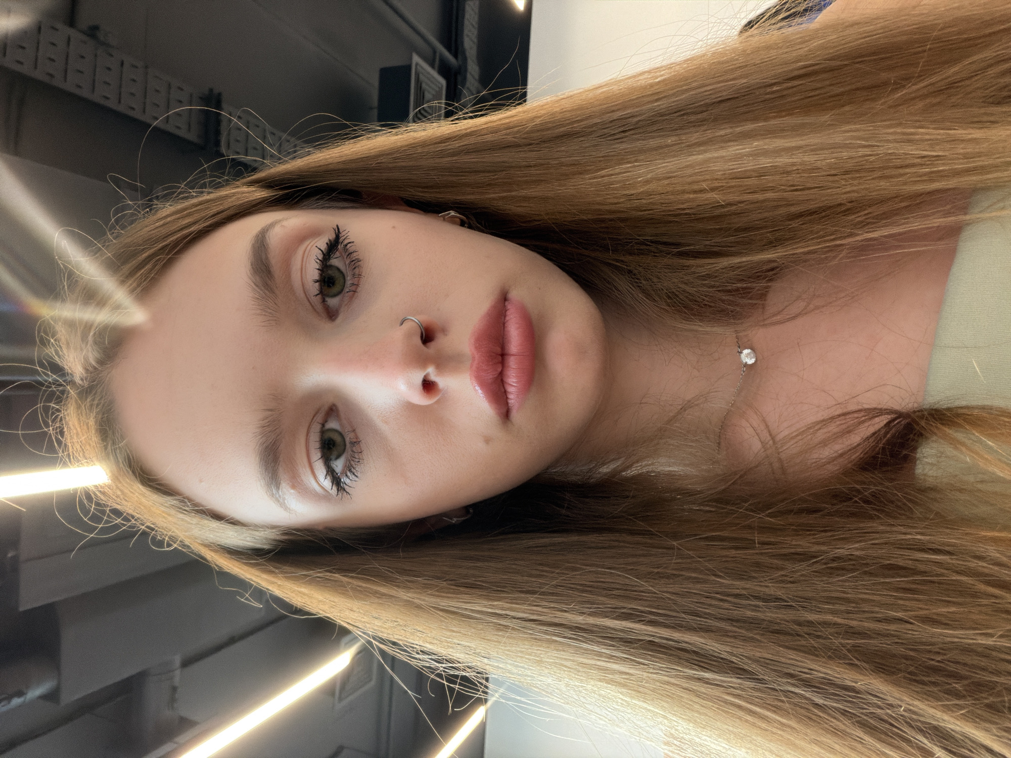 A close-up, head-on, outdoor, medium shot shows a young woman with long, light brown hair. Her hair is parted in the middle and falls down past her shoulders. She is looking directly at the camera with a neutral expression. The woman is wearing makeup, including mascara and lipstick. She has a nose ring and small hoop earrings. A thin necklace with a diamond pendant is visible around her neck. The background is blurred, but appears to be an indoor setting with overhead lighting and dark ceiling panels.